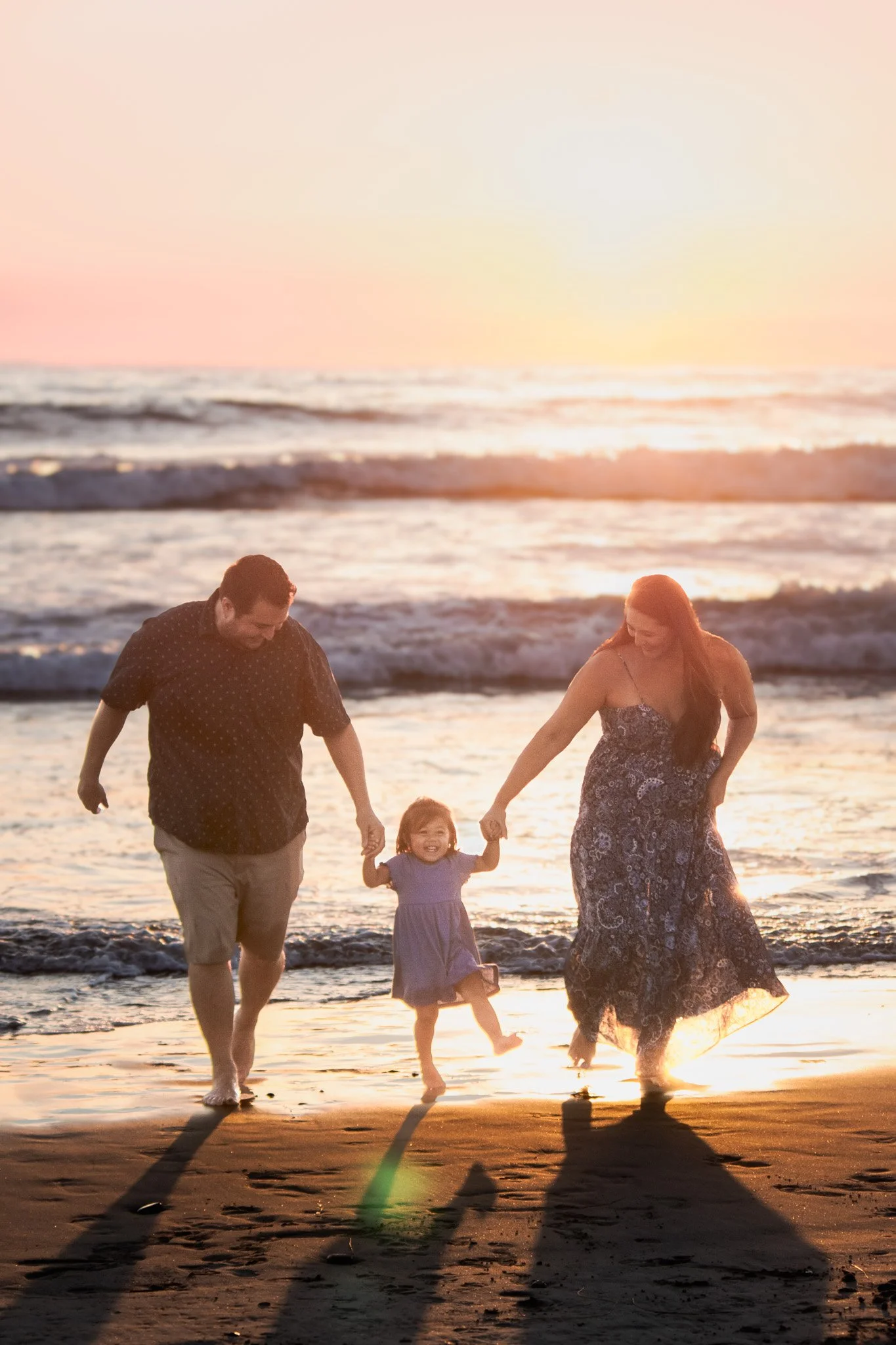 A family of four walking hand in hand along the beach at sunset, with the ocean waves in the background and their shadows stretching on the sand.