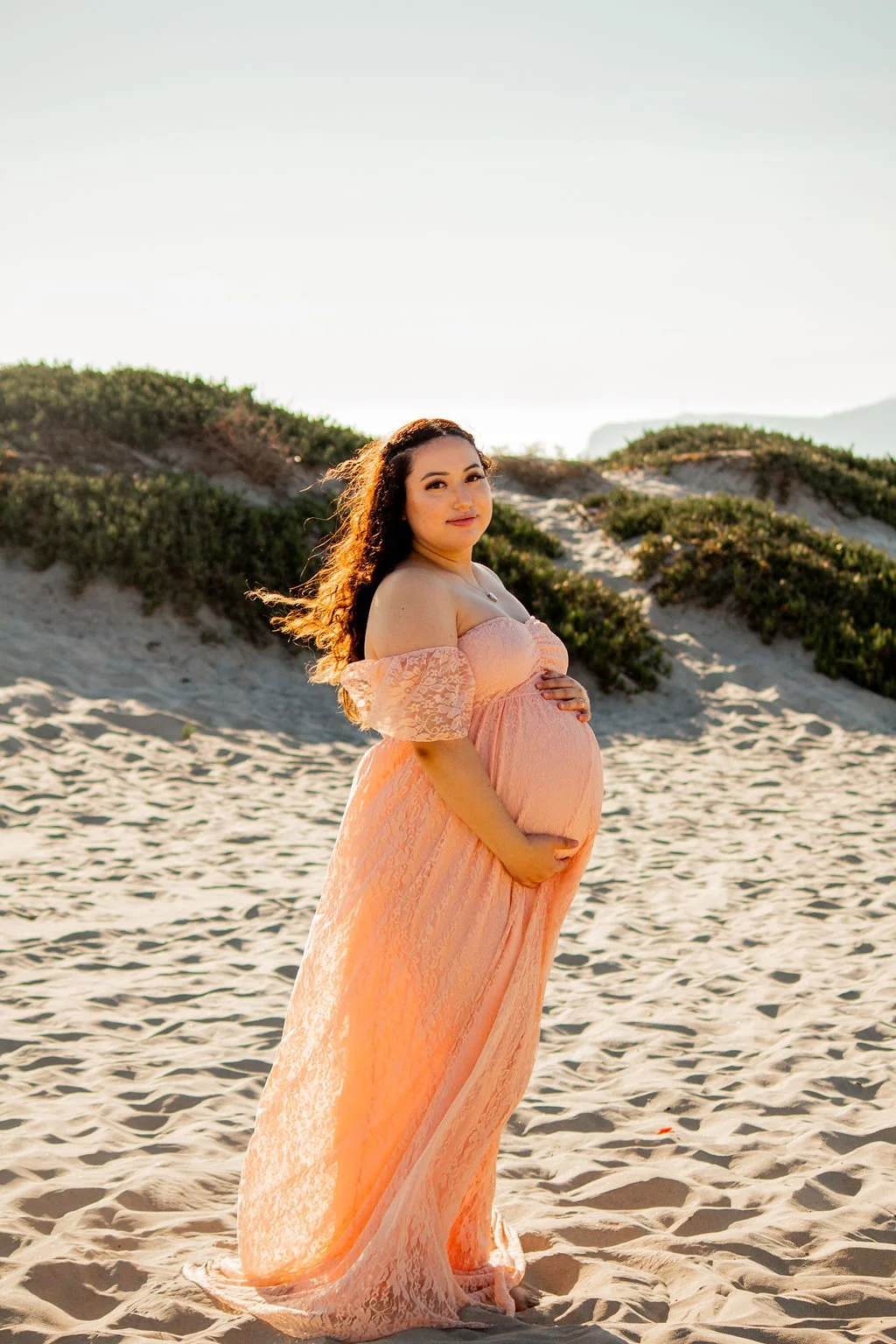 A pregnant woman in a peach lace off-the-shoulder dress standing on a sandy beach with green dunes in the background, during sunset.