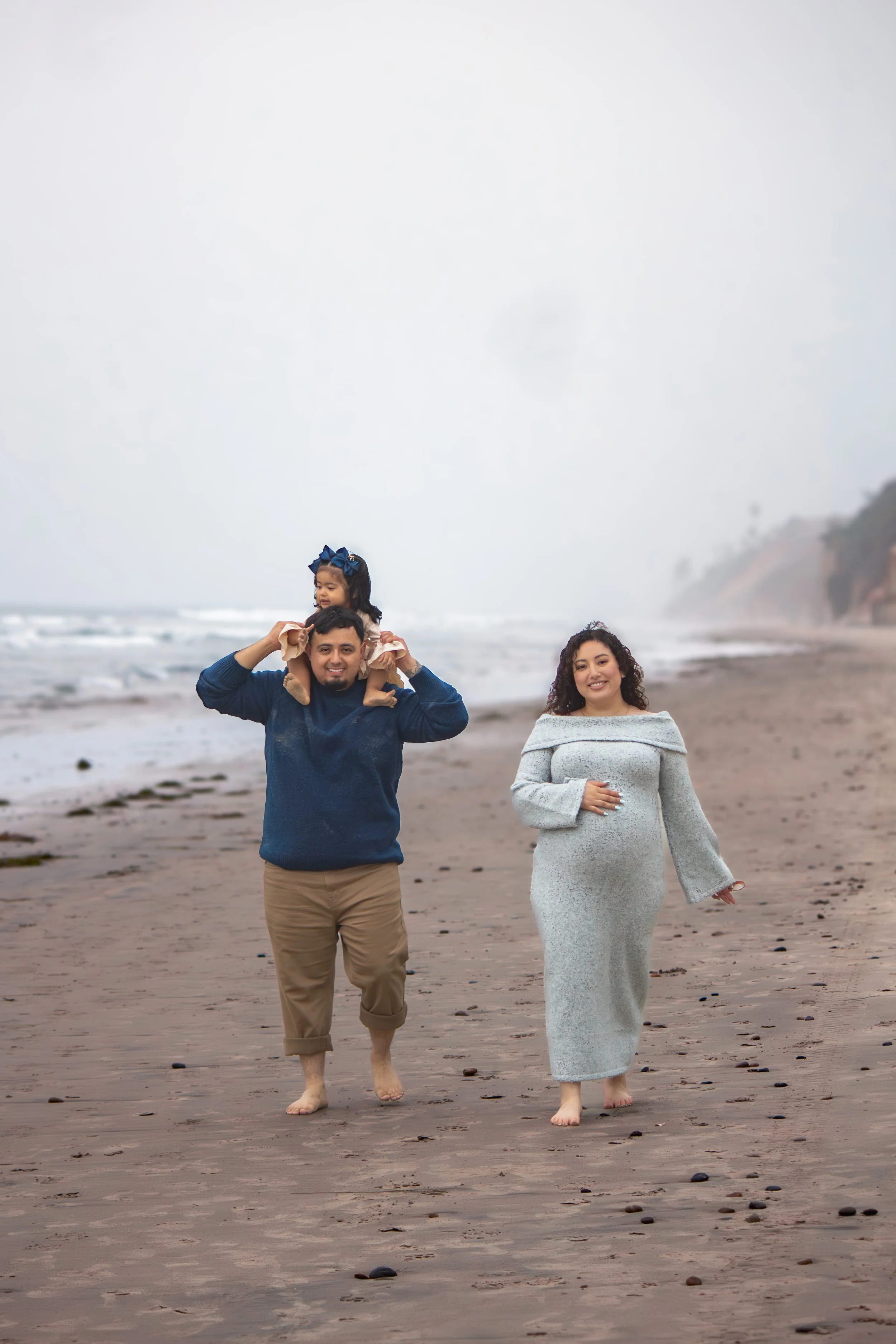 Family walking on the beach, with a man carrying a young girl on his shoulders and a woman walking beside them, ocean and cloudy sky in the background.