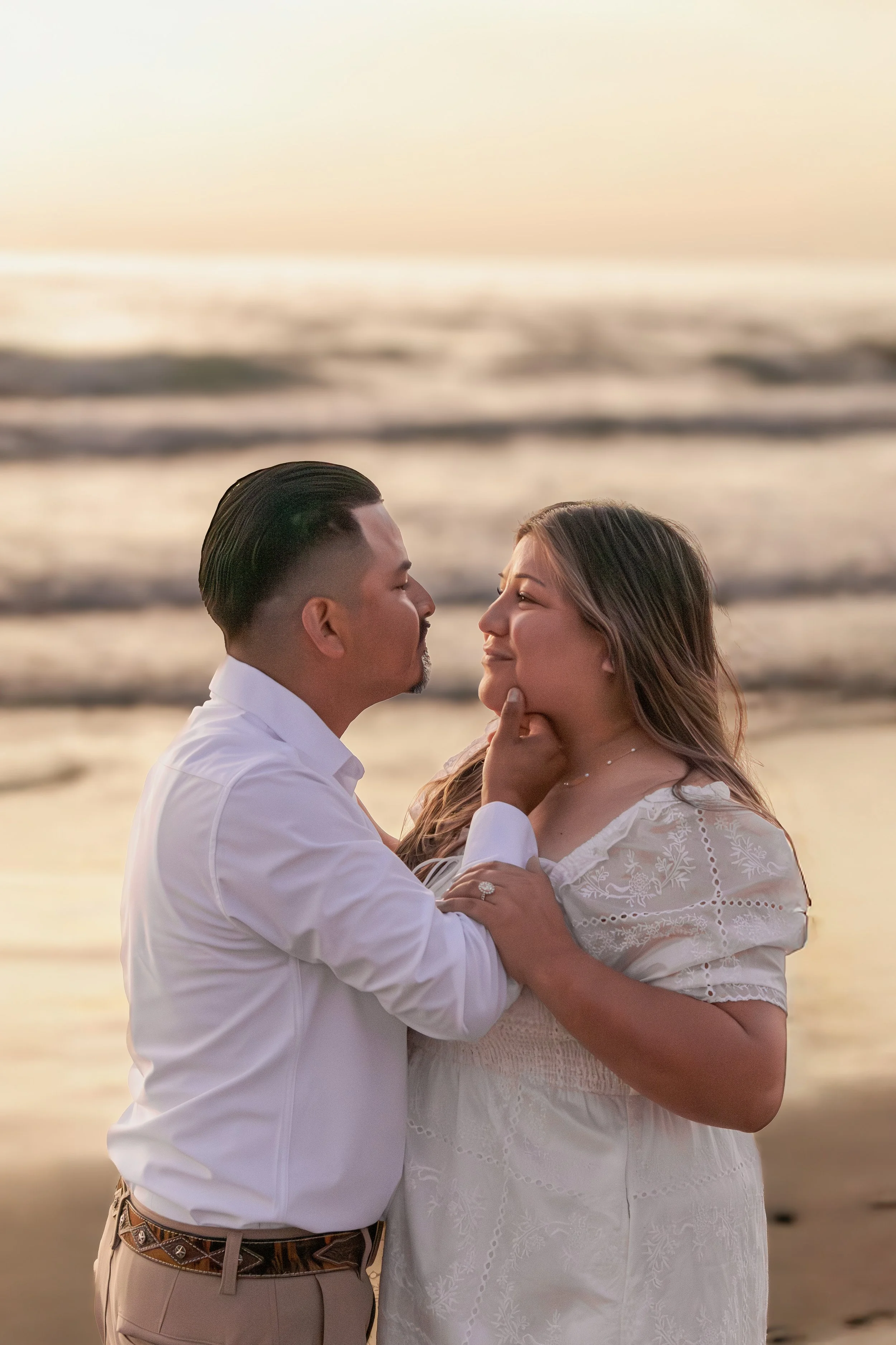 A couple stands close together on a beach during sunset, gazing into each other's eyes, with the ocean waves in the background.