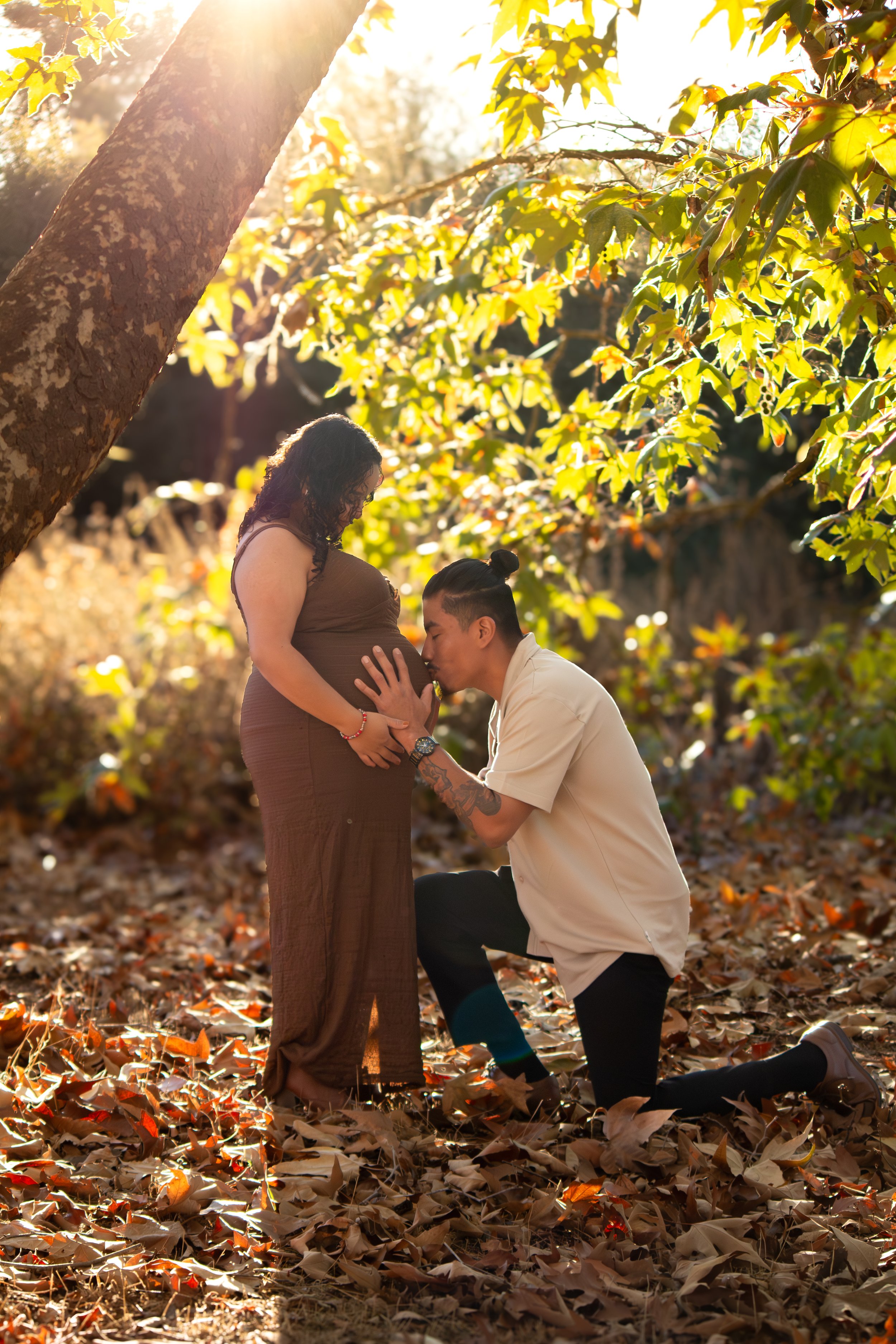 A man kneeling and kissing the pregnant belly of a woman standing outdoors amidst fall leaves, sun shining through trees.