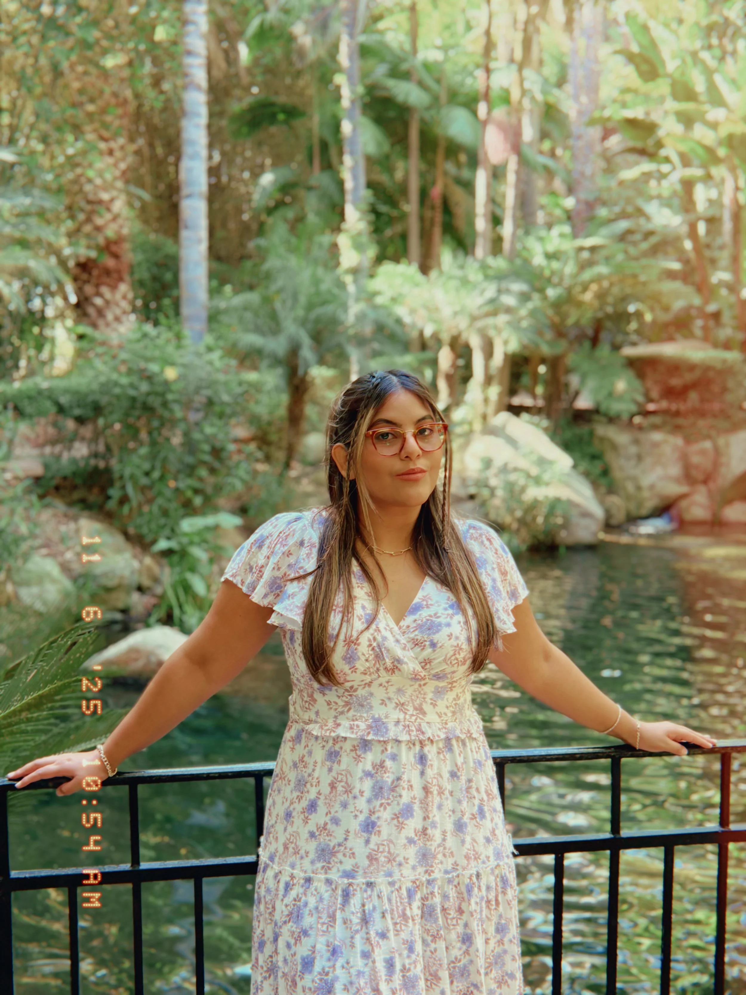 A woman standing outdoors near a railing by a pond or stream in a lush, green forest with tall trees and sunlight filtering through the leaves.