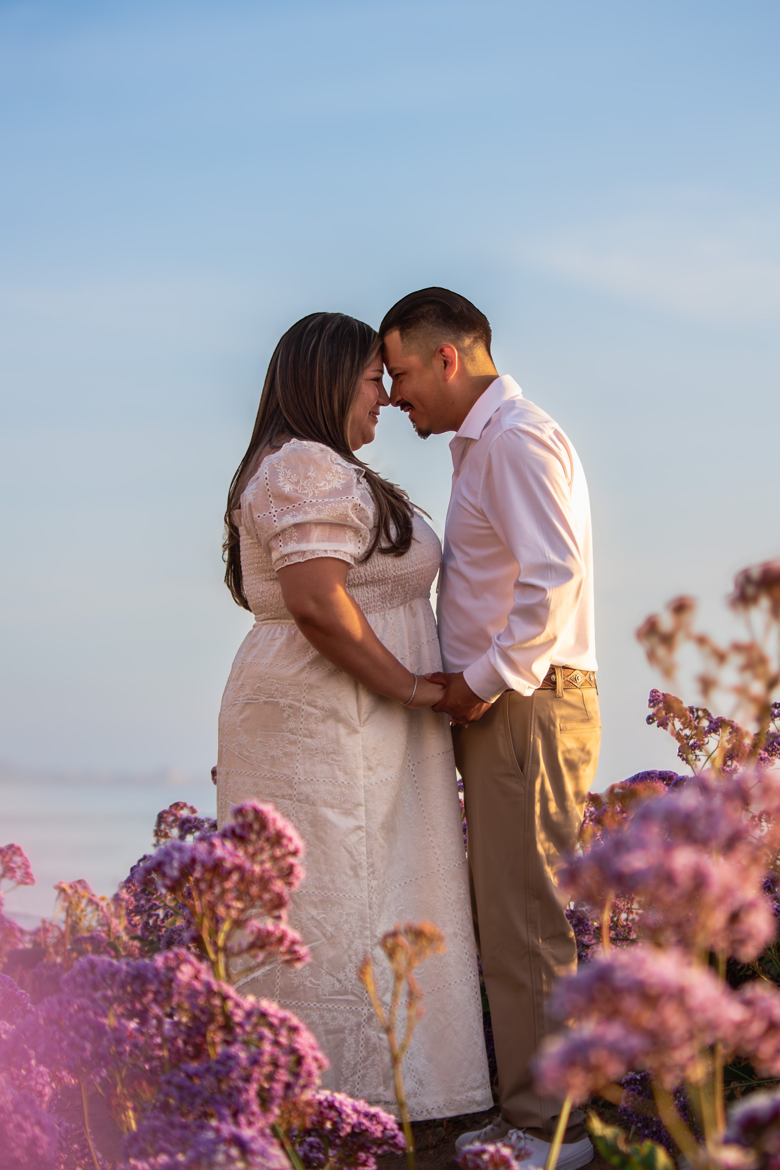 A couple stands close together with foreheads touching, holding hands on a beach with pink and purple flowers in the foreground during sunset.