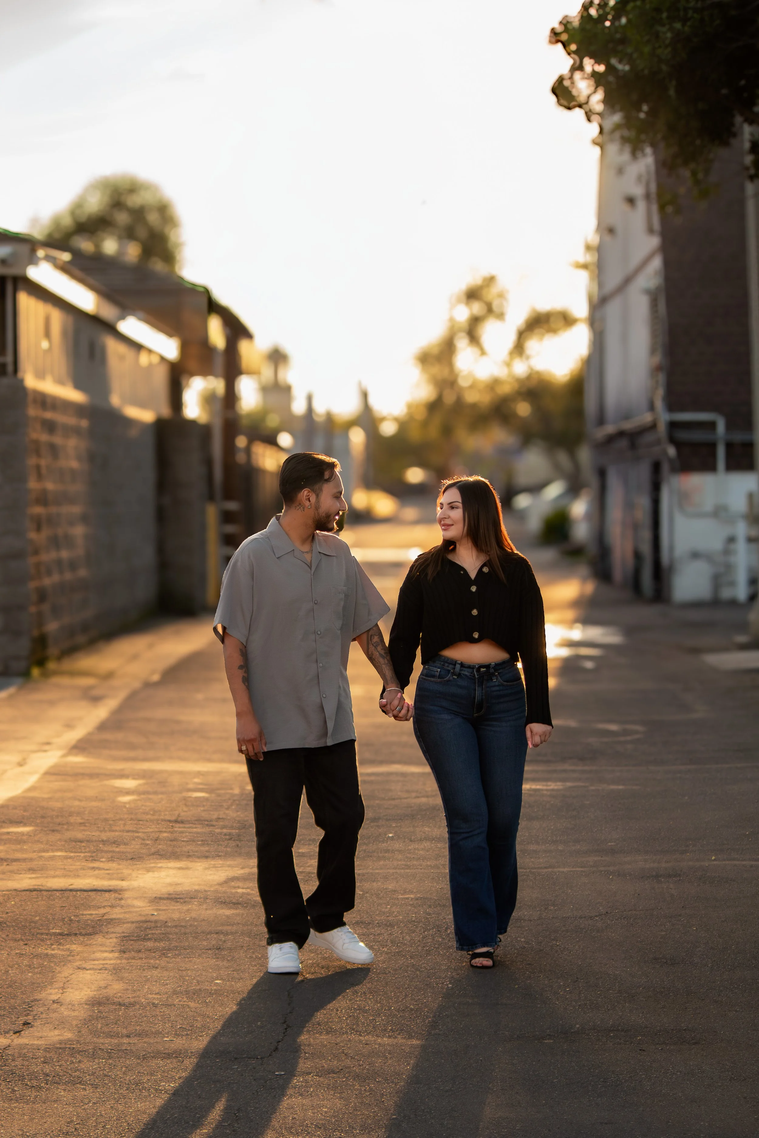 A young man and woman walk hand-in-hand on a street during sunset, smiling at each other, with houses and trees in the background.
