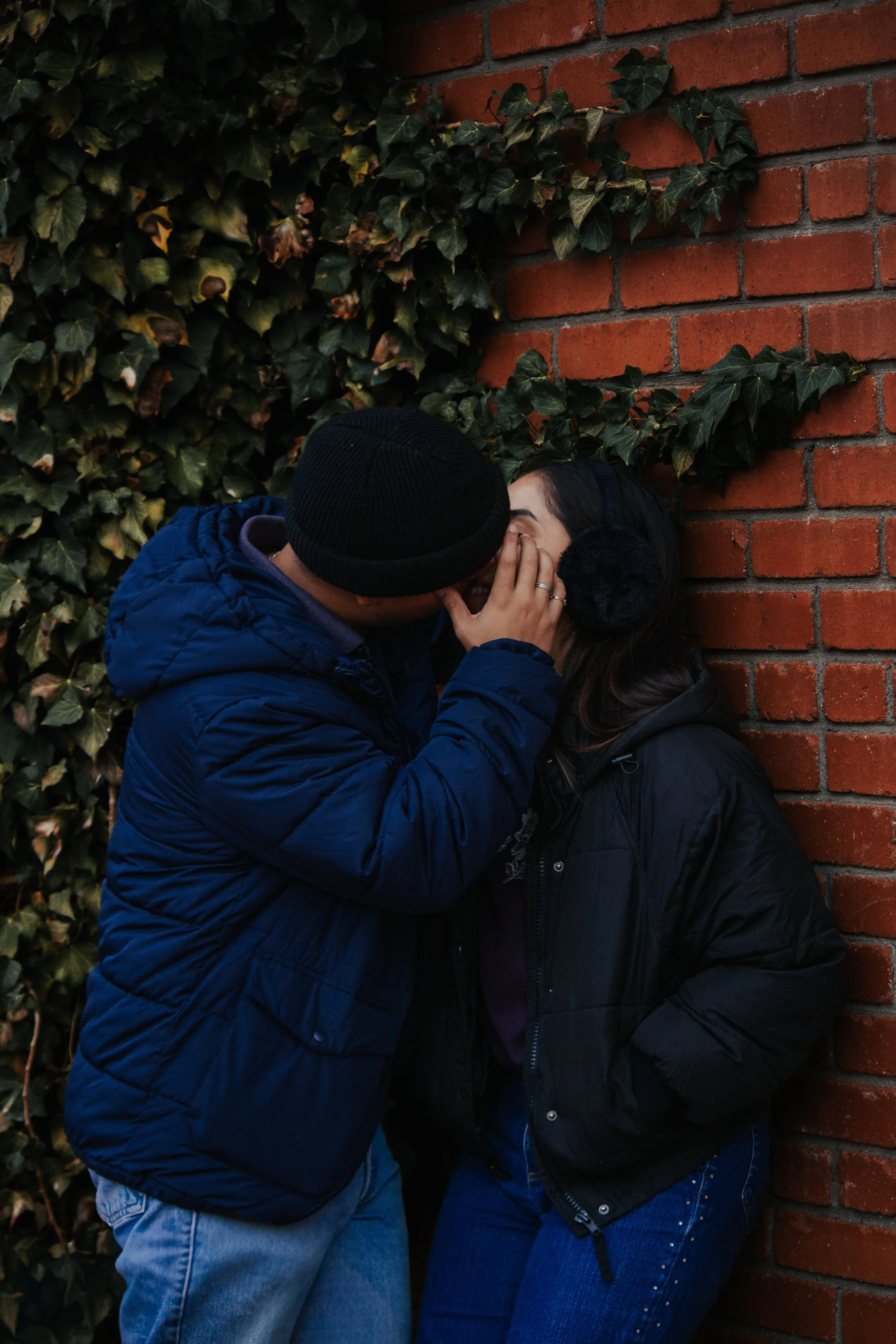 A couple sharing a kiss against a brick wall with green ivy leaves. The man is wearing a black beanie and blue jacket, and the woman has dark hair and is wearing a black jacket.