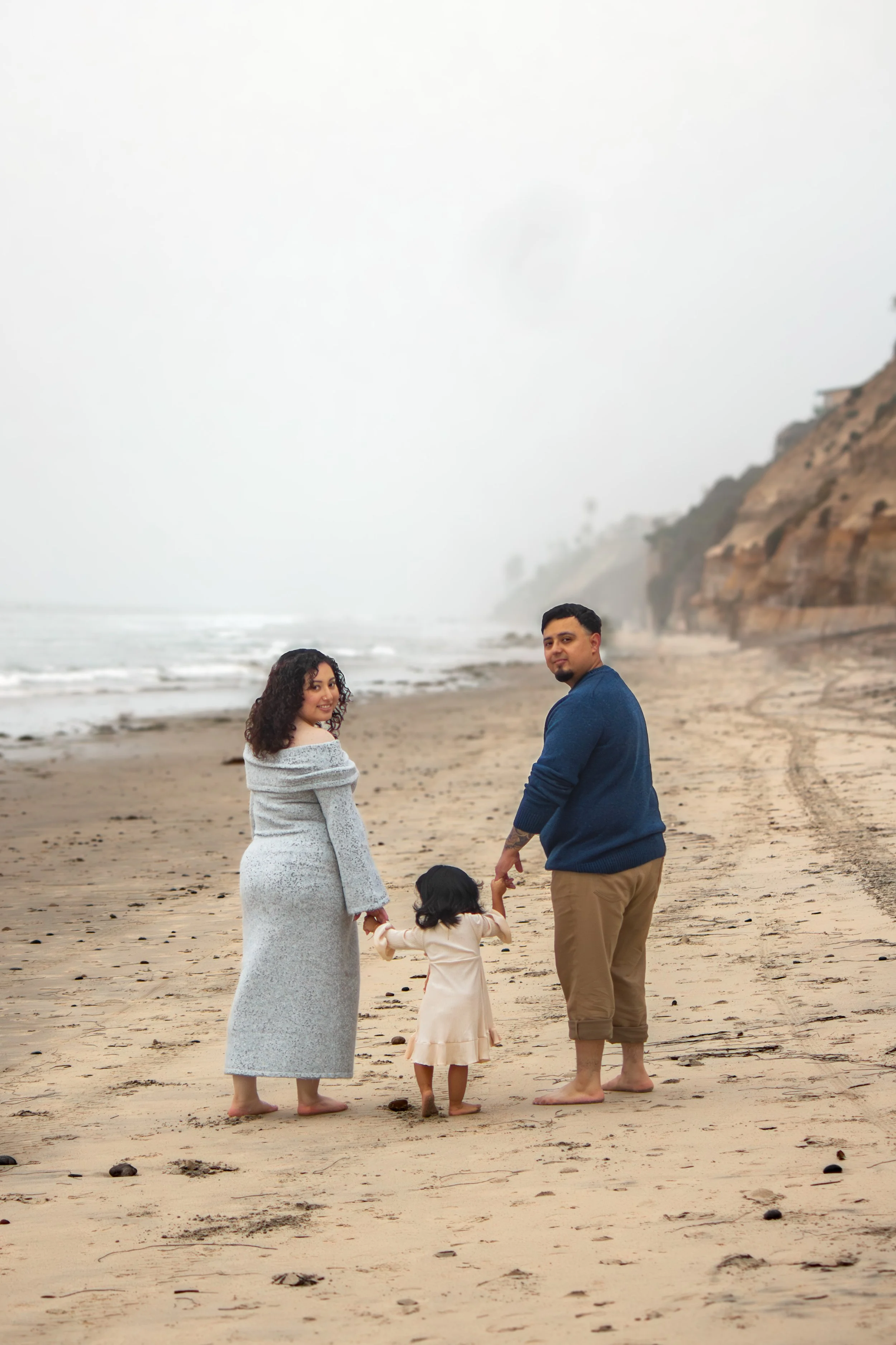 A family on a beach during overcast weather. The woman is wearing a long gray dress, and the man is wearing khaki pants and a blue sweater. A young girl in a beige dress holds hands with an adult woman, while the woman holds the child's hand with her left hand. The ocean waves are visible in the background, along with rocky cliffs.
