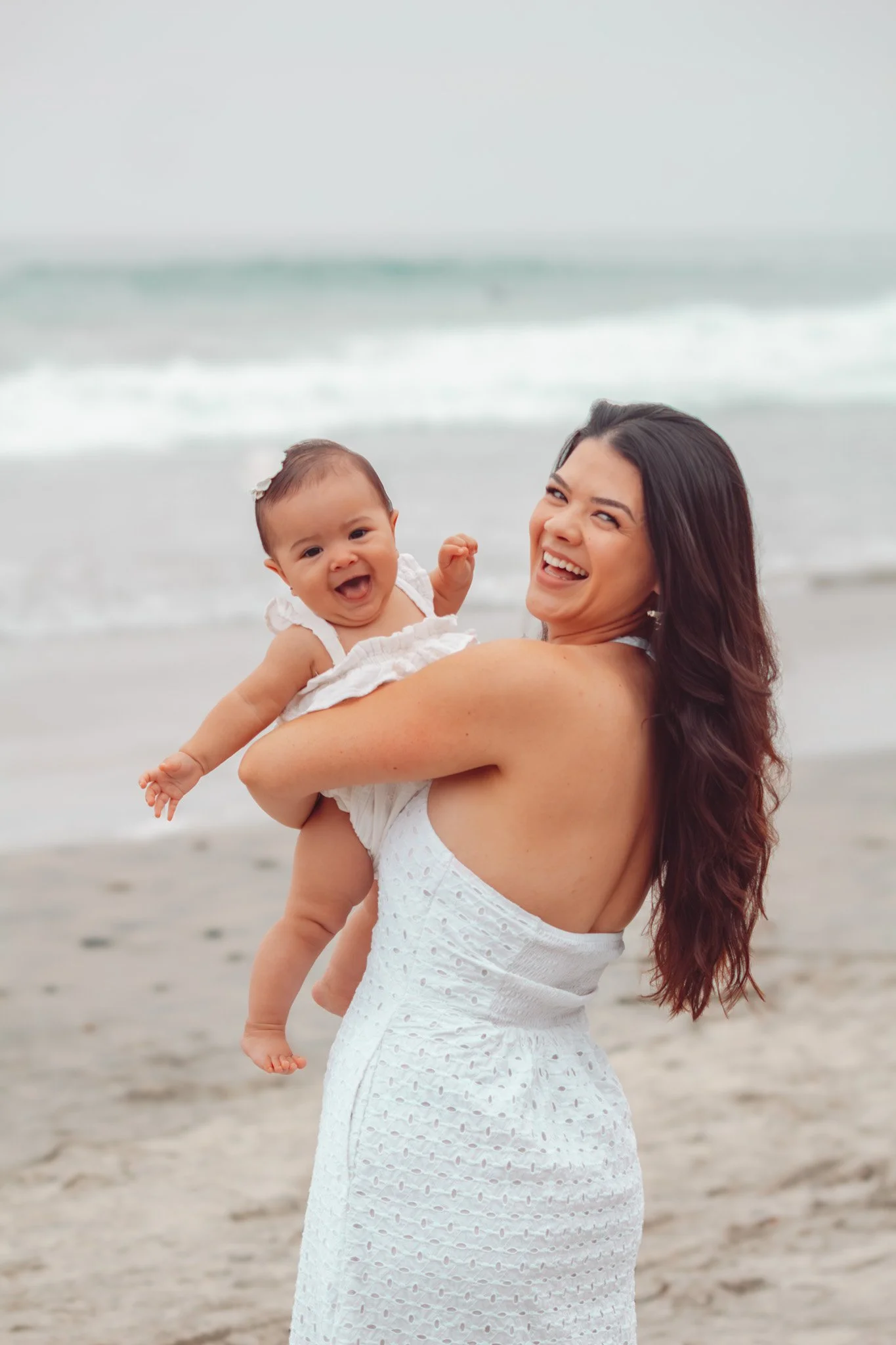 A woman with long, wavy brown hair wearing a white dress, holding a smiling baby girl in a white dress at the beach, with waves and ocean in the background.