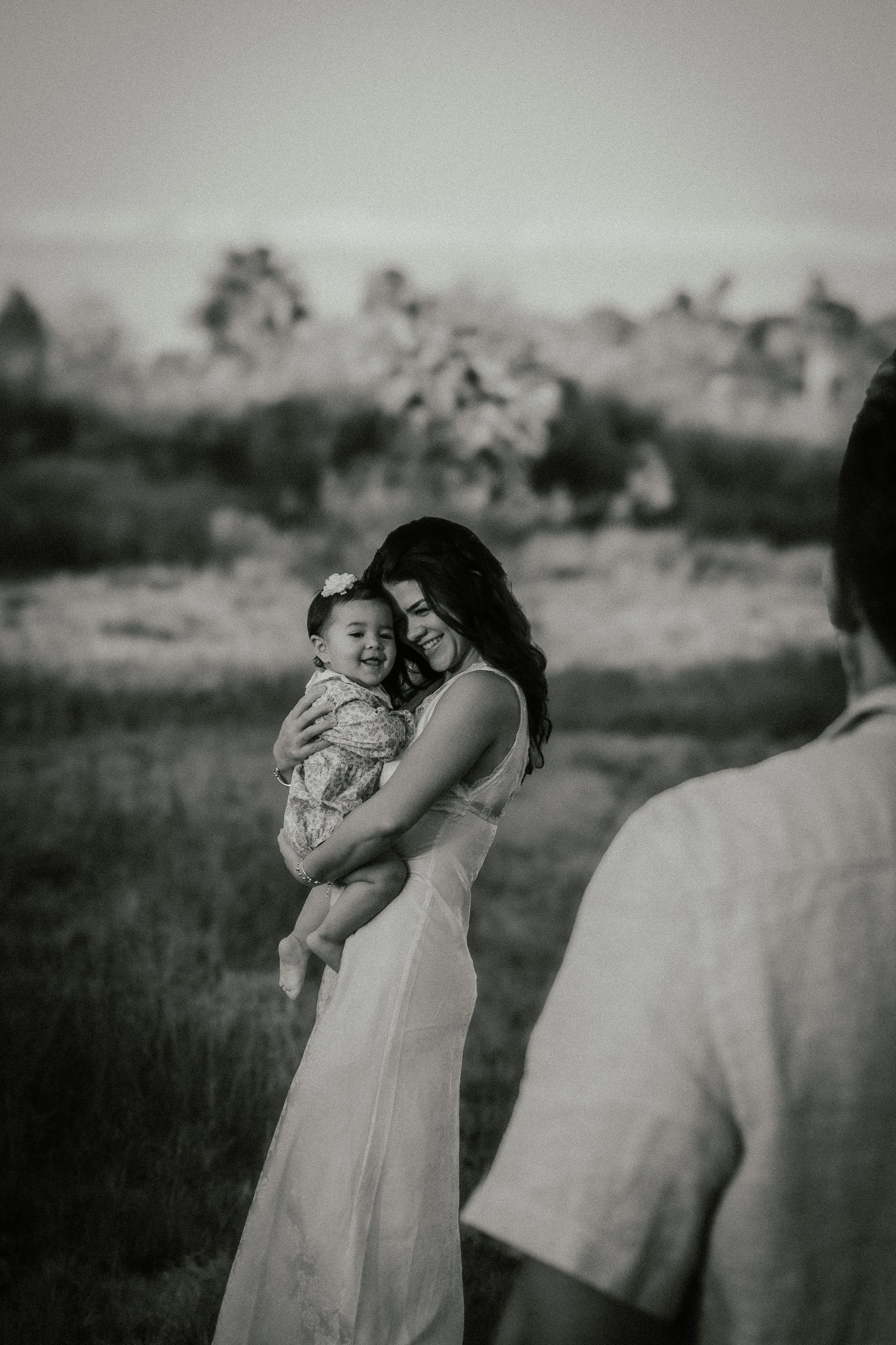 A woman in a white dress is holding a smiling young girl in a floral dress and a headband. They are outdoors in a field with trees in the background, and the photo is in black and white.