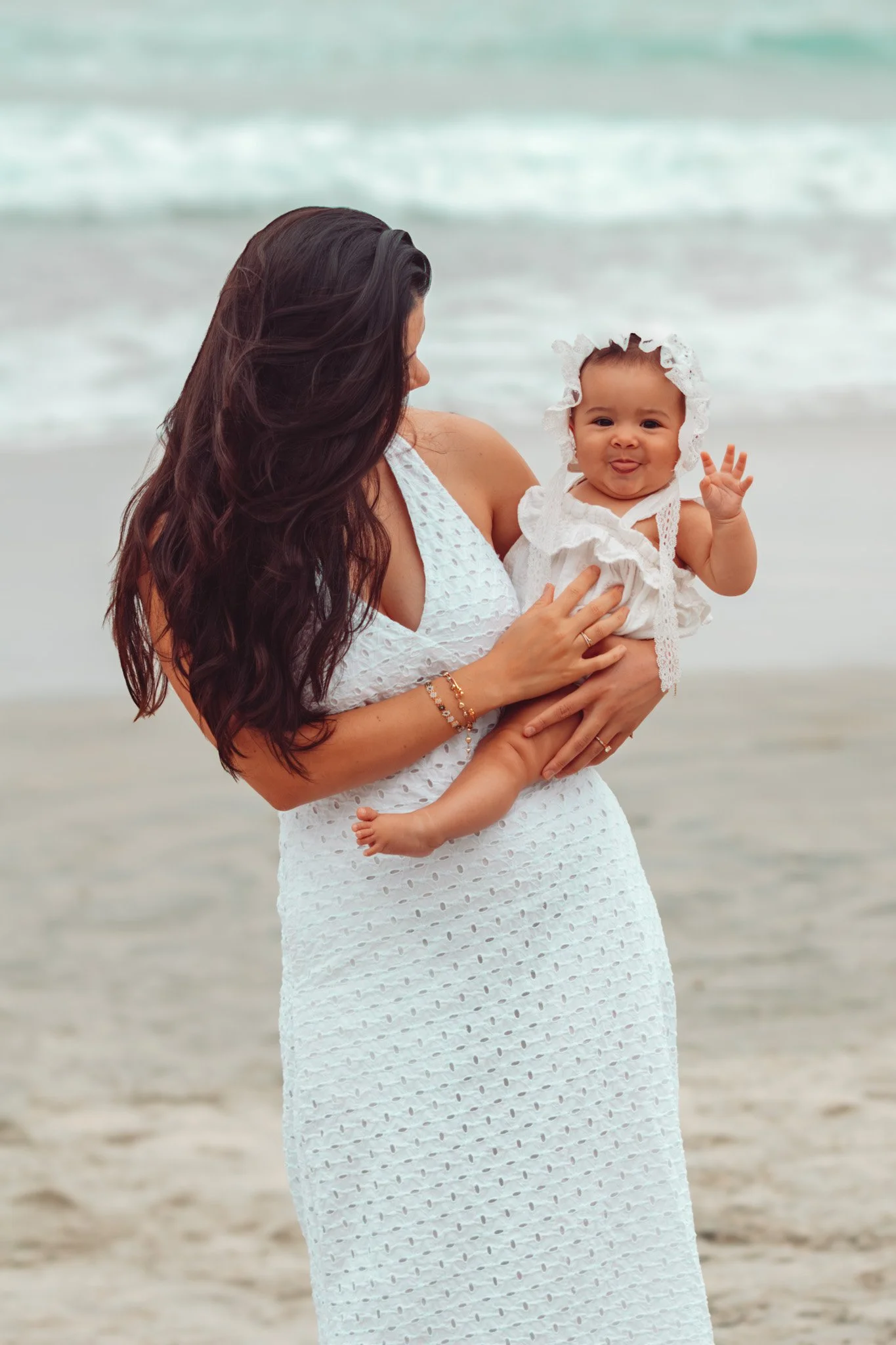 A woman in a white dress holding a baby girl at the beach with waves in the background.