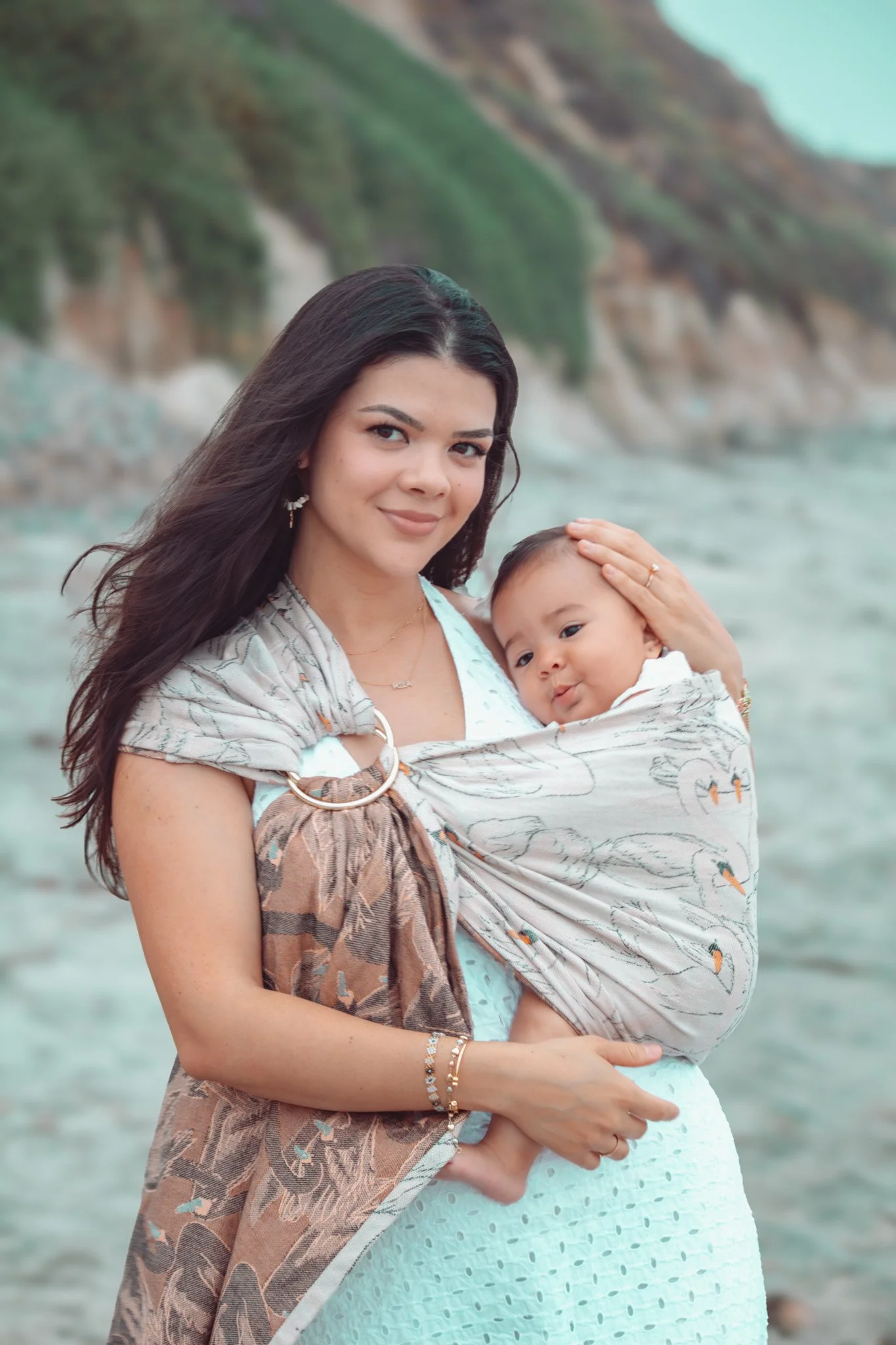 A woman holding a baby on beach with cliffs and ocean in background.