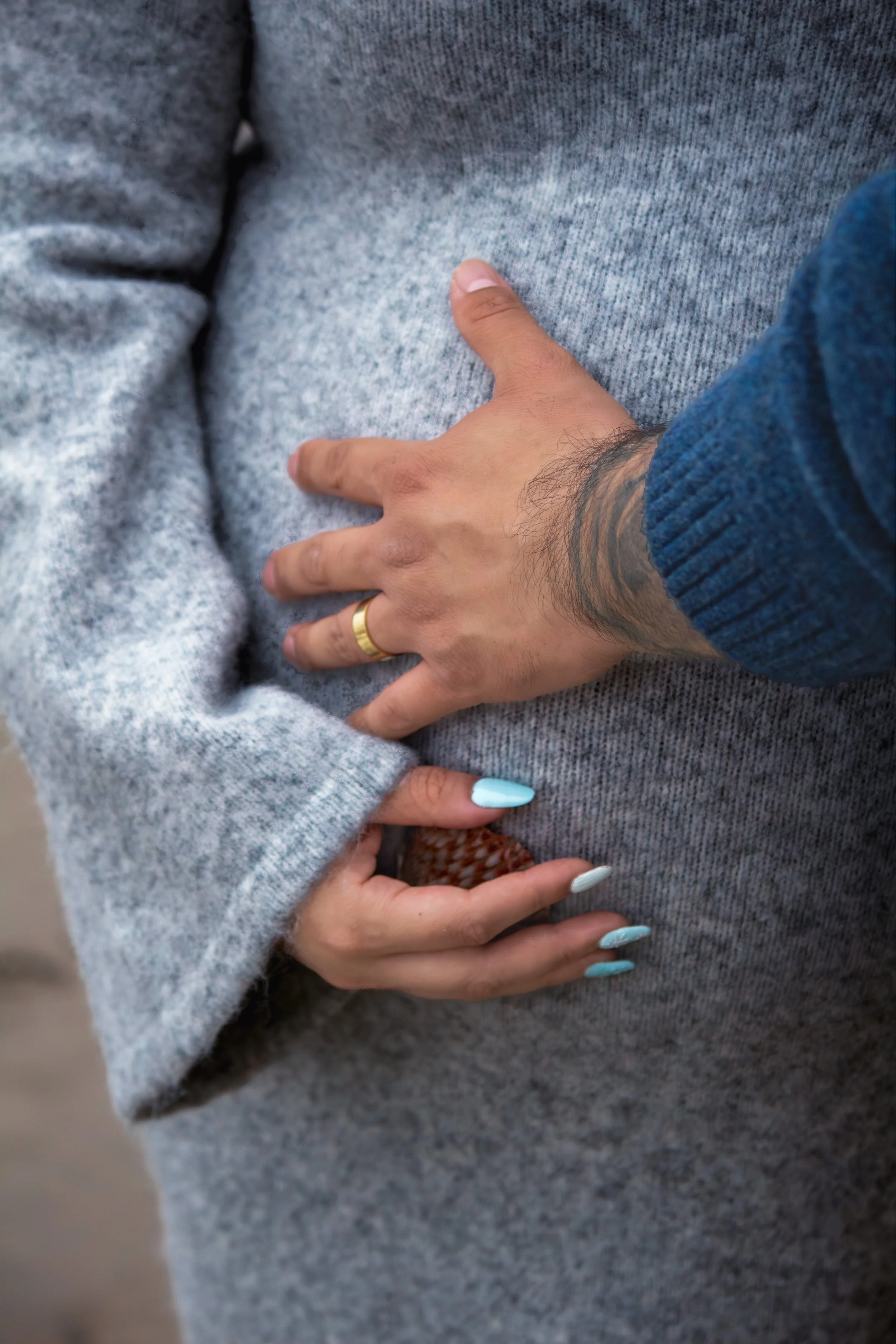 A person's hand with a wedding ring resting on gray fabric, with another hand below, featuring manicured nails painted light blue. The person is wearing a blue sweater and has a tattoo on the wrist.
