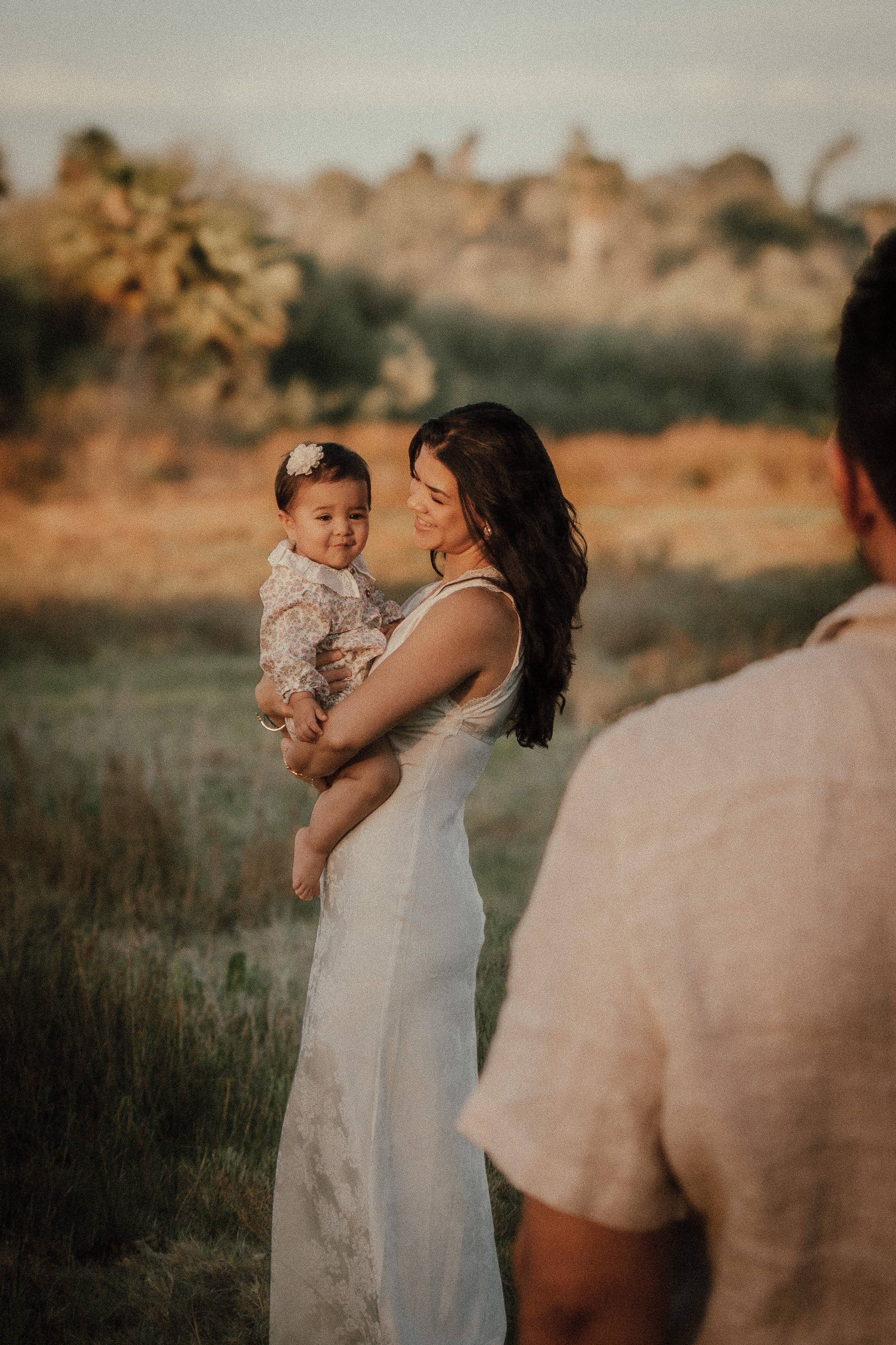 A woman in a white dress holding a young girl with a flower headband at the beach during sunset, with a blurry background of trees and the ocean, and part of a person in the foreground.