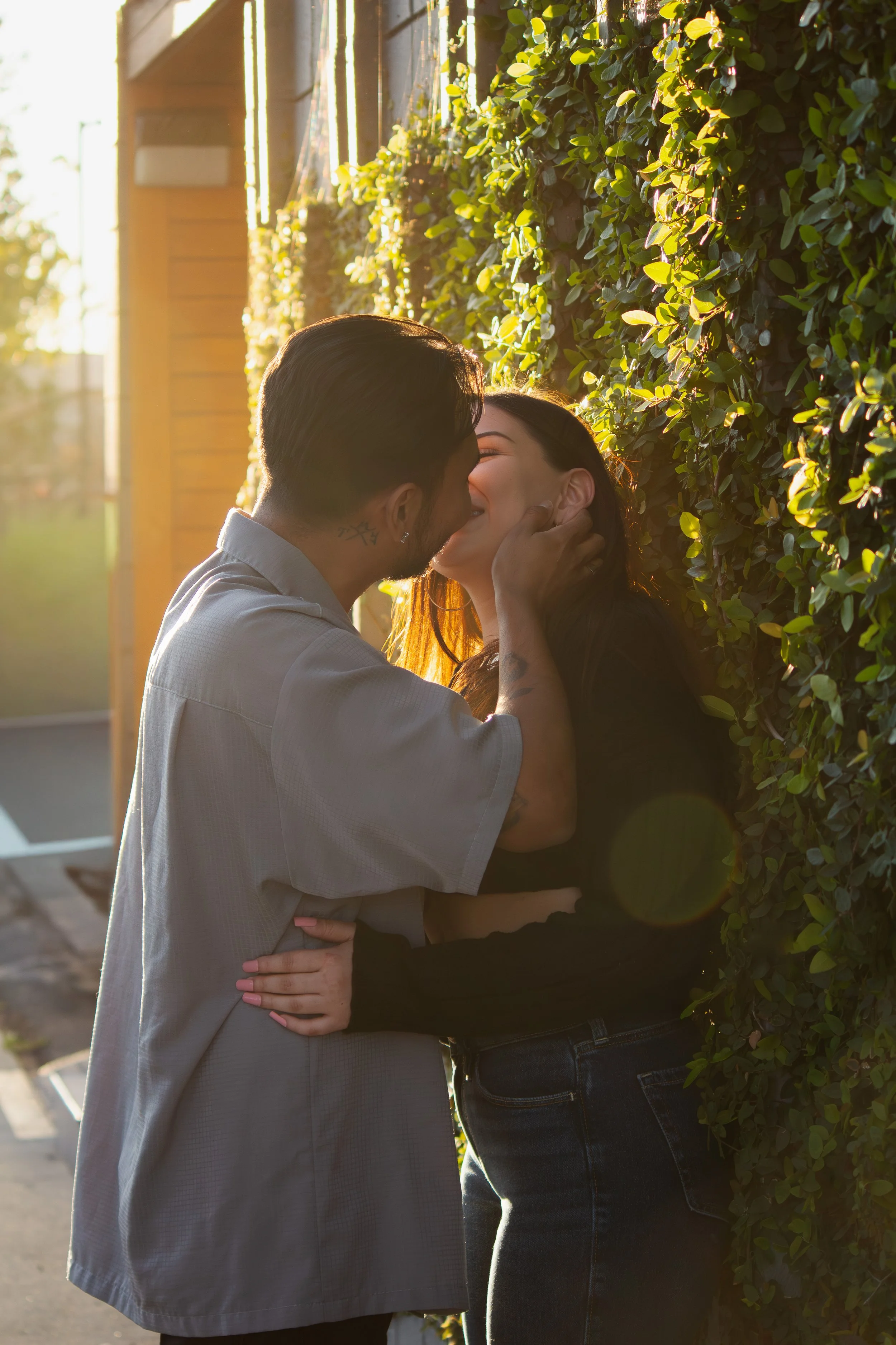 A couple sharing a kiss outdoors in front of a leafy hedge during sunset, with their faces close and smiles, creating a romantic scene.
