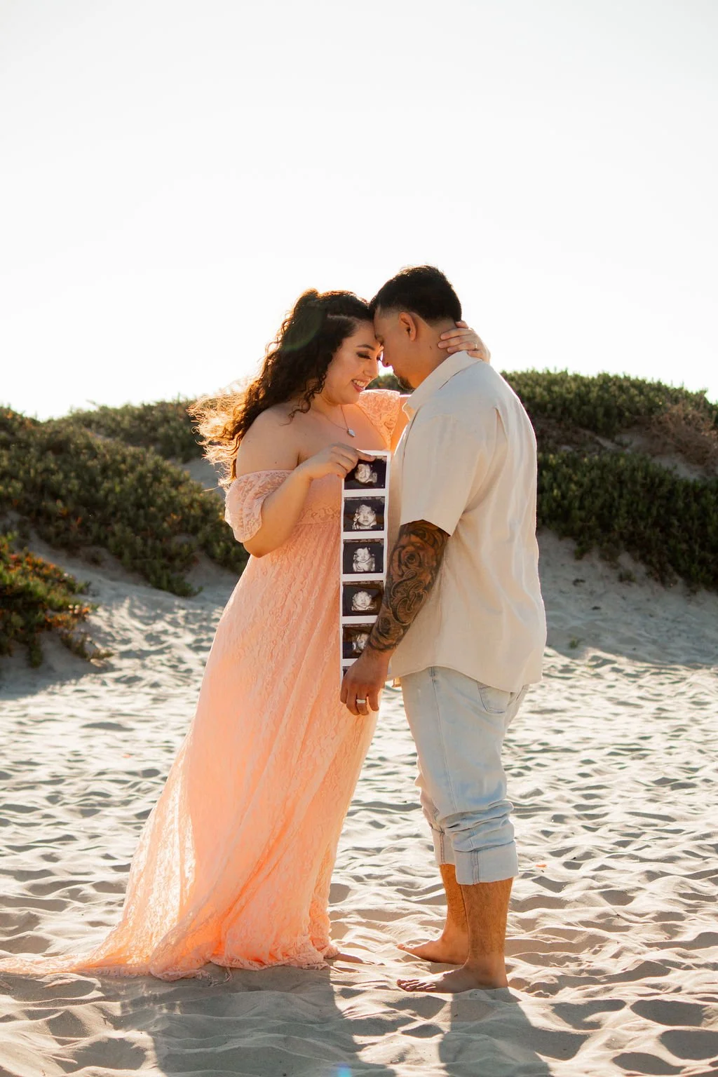 A couple on a beach holding ultrasound images, smiling, with their foreheads touching, during sunset.