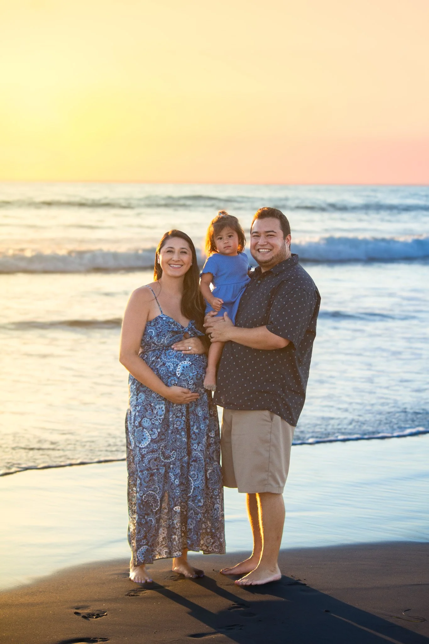 A family of three, consisting of a woman, a man, and a young girl, standing on a beach at sunset. The woman is wearing a blue patterned dress, the man is wearing a navy blue shirt with white dots and khaki shorts, and the girl is in a blue dress. The