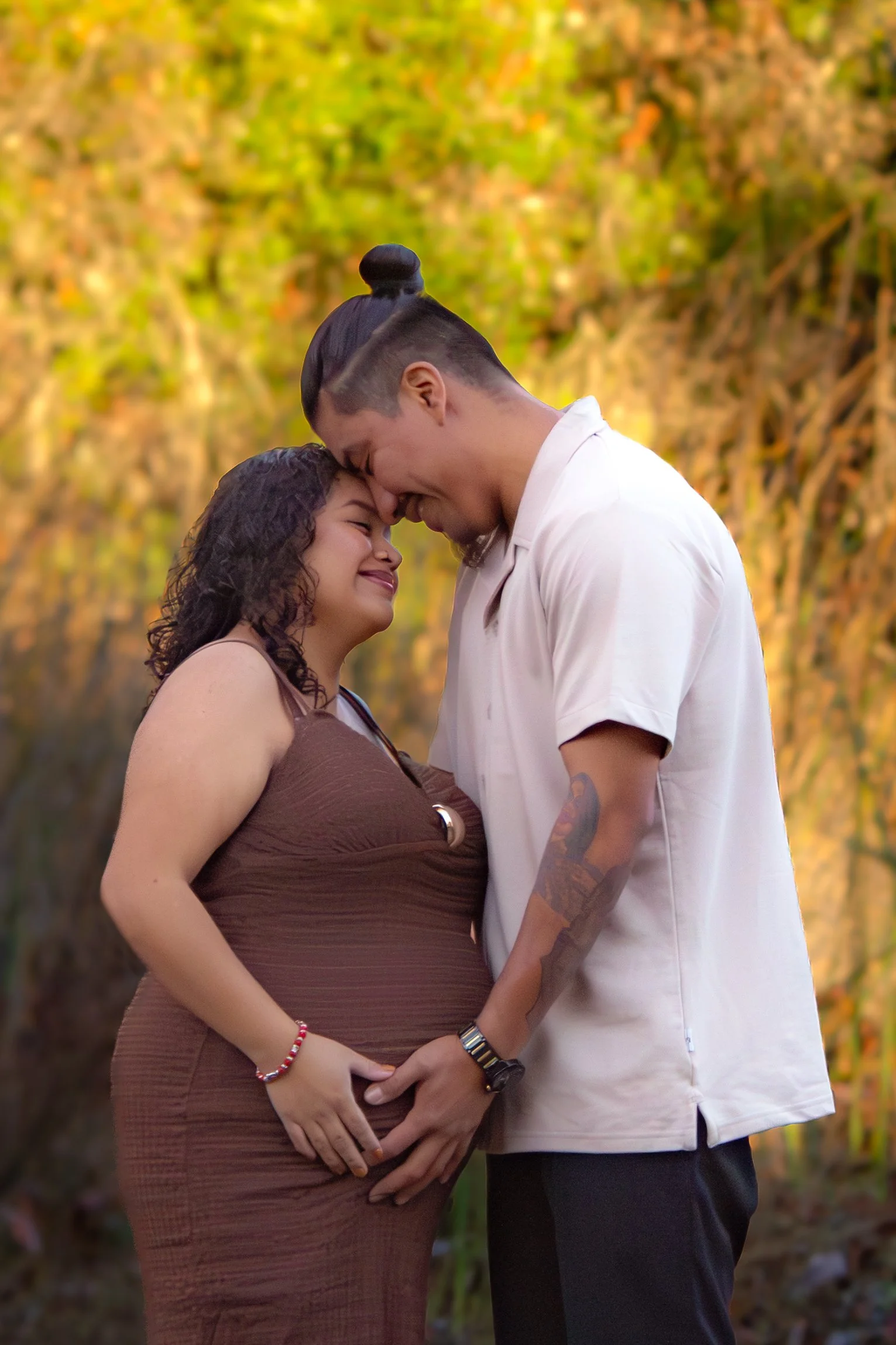 A pregnant woman and man stand outdoors with their foreheads touching, smiling, in front of a tree with autumn leaves.