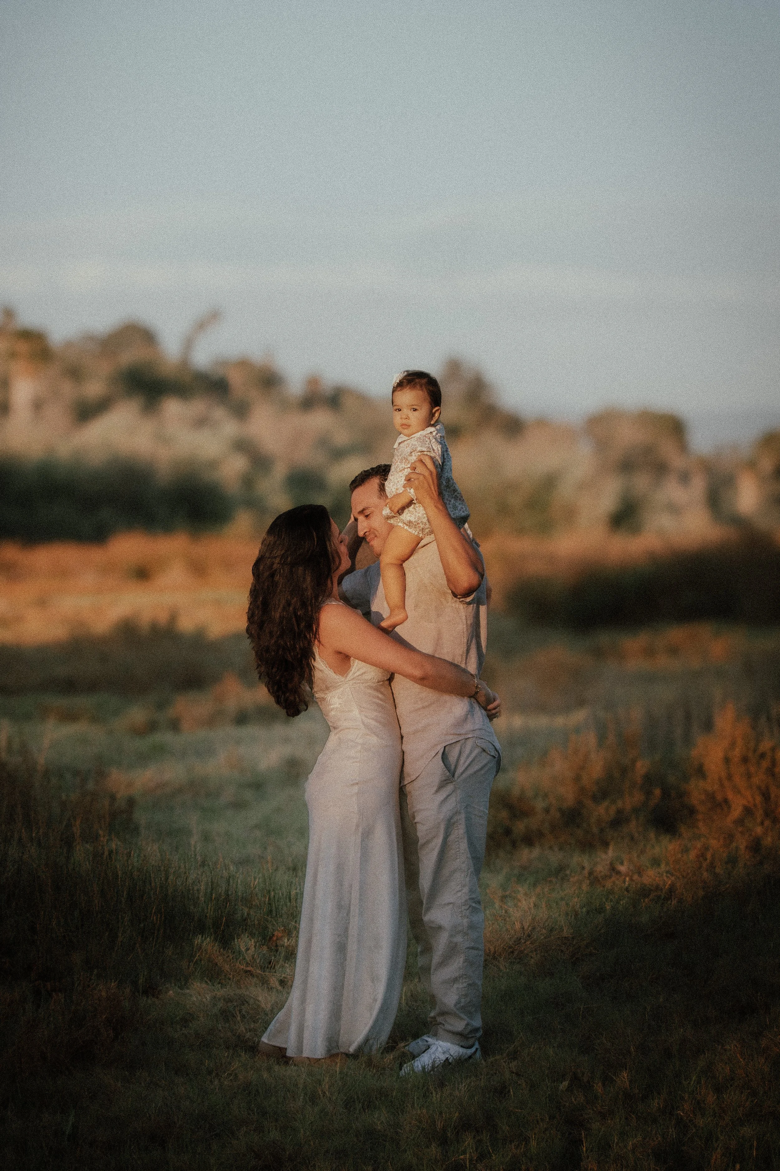 A family of three, a man, a woman, and a young child, standing outdoors in a grassy area with hills in the background during sunset, enjoying a moment together.