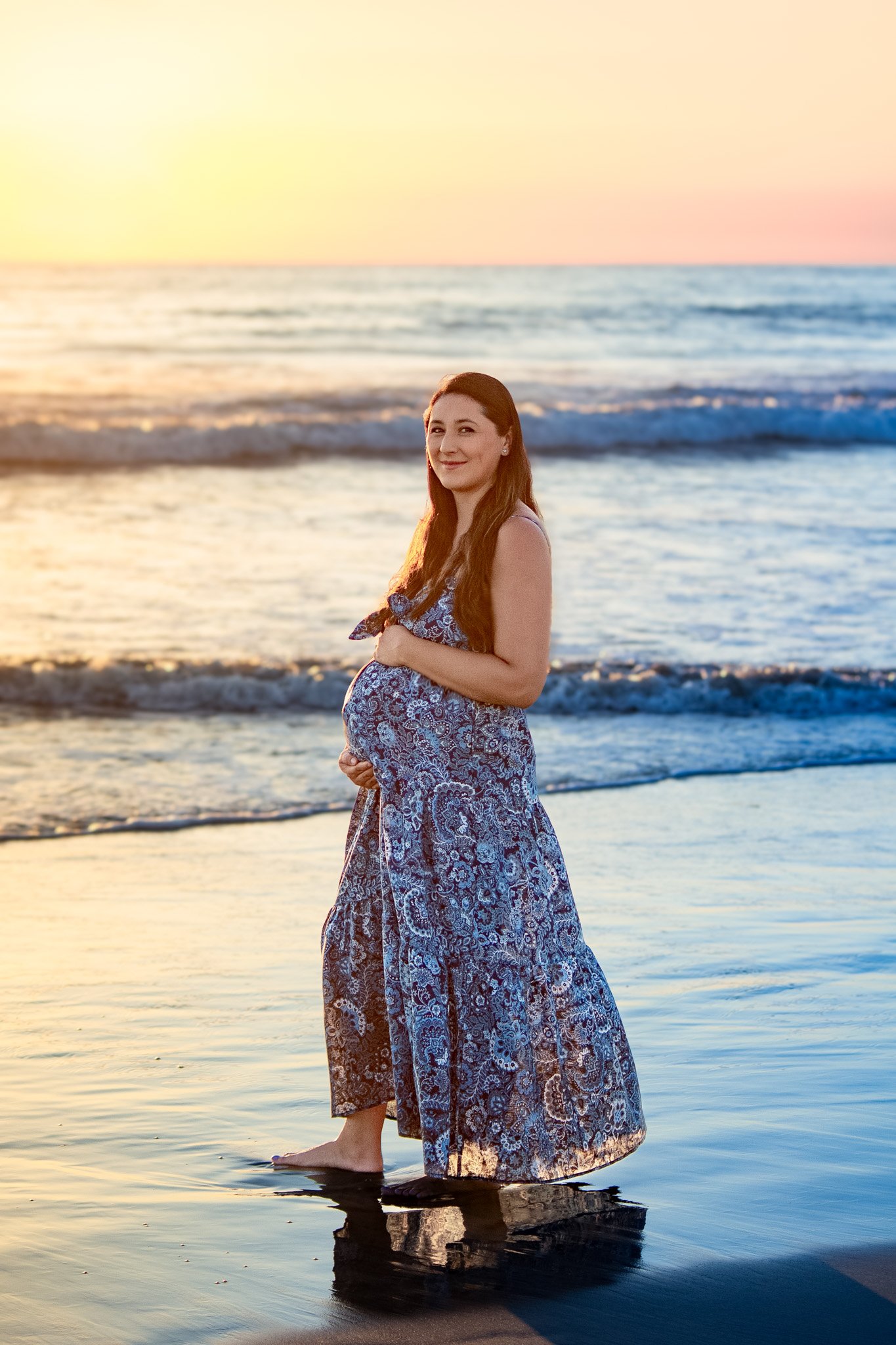 A pregnant woman standing in shallow ocean water during sunset, wearing a blue patterned maxi dress, smiling at the camera.