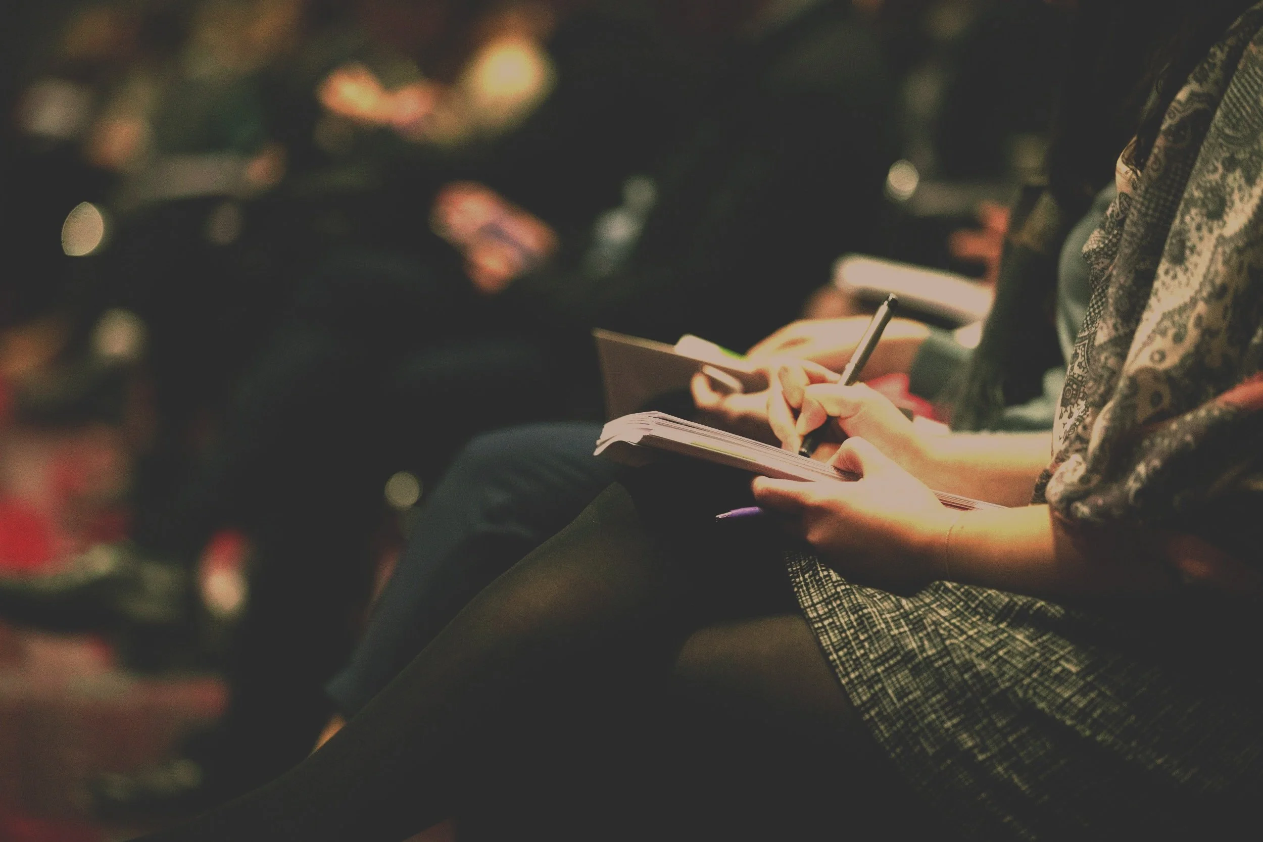Person sitting in a dark setting taking notes or writing in a notebook with a pen, wearing patterned clothing.