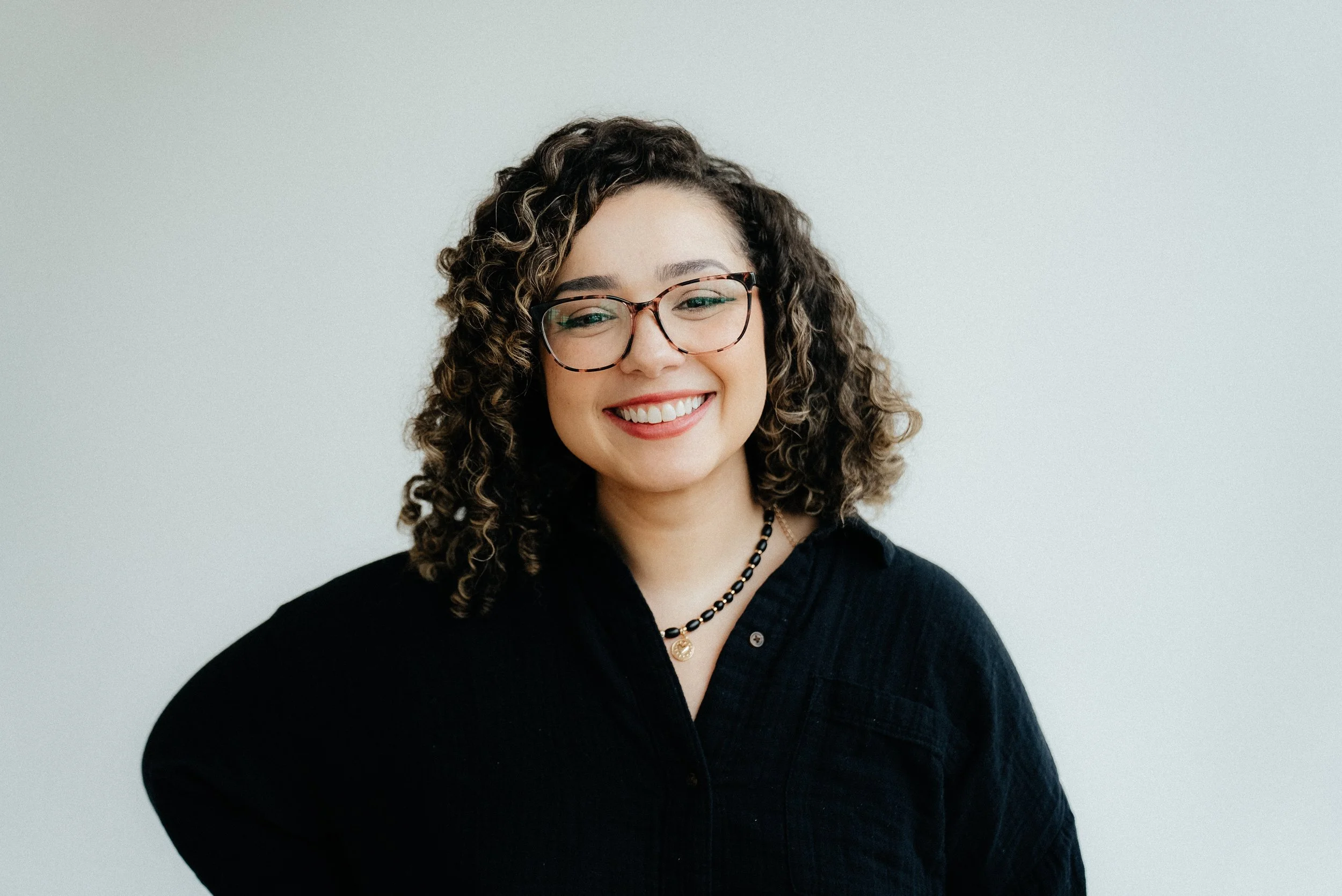 A young woman with curly hair, wearing glasses, a black top, and jewelry, smiling against a plain light gray background.