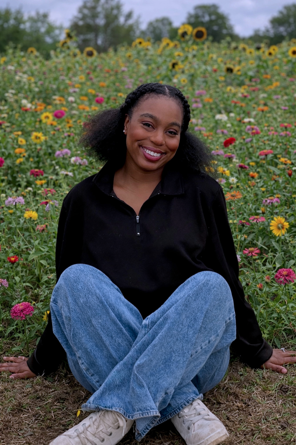 A smiling woman with curly hair sitting on the ground in a flower field with yellow, pink, orange, and white flowers, wearing a black jacket, blue jeans, and white sneakers.