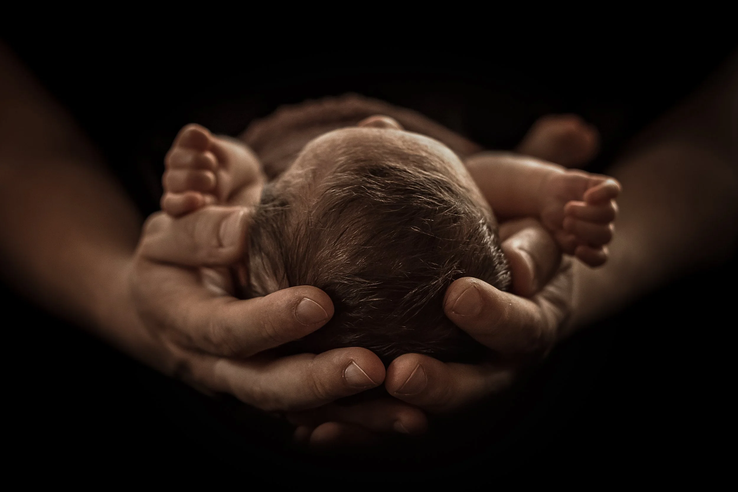 Hands gently cradle a newborn baby with closed eyes, dark background.