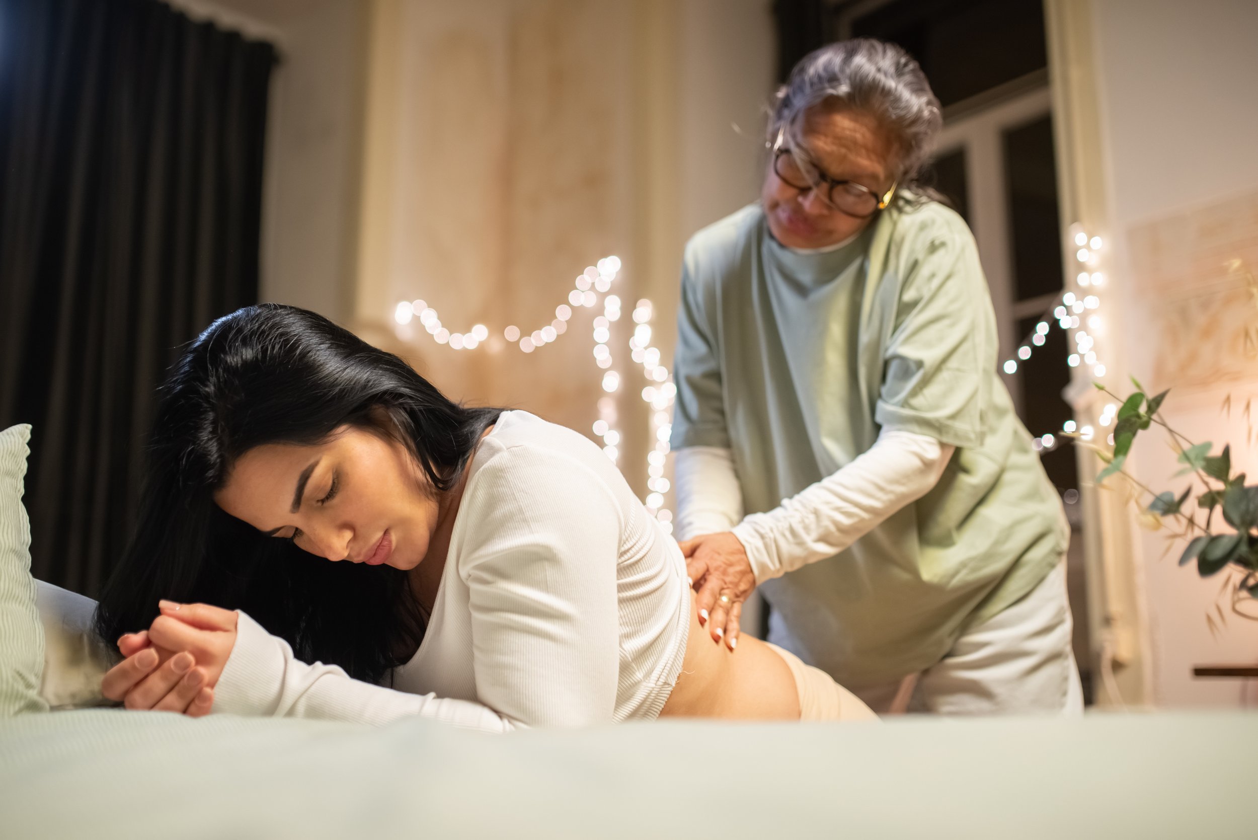 A woman laying on a bed with her eyes closed, receiving a massage from an older woman in a room decorated with string lights and a potted plant.