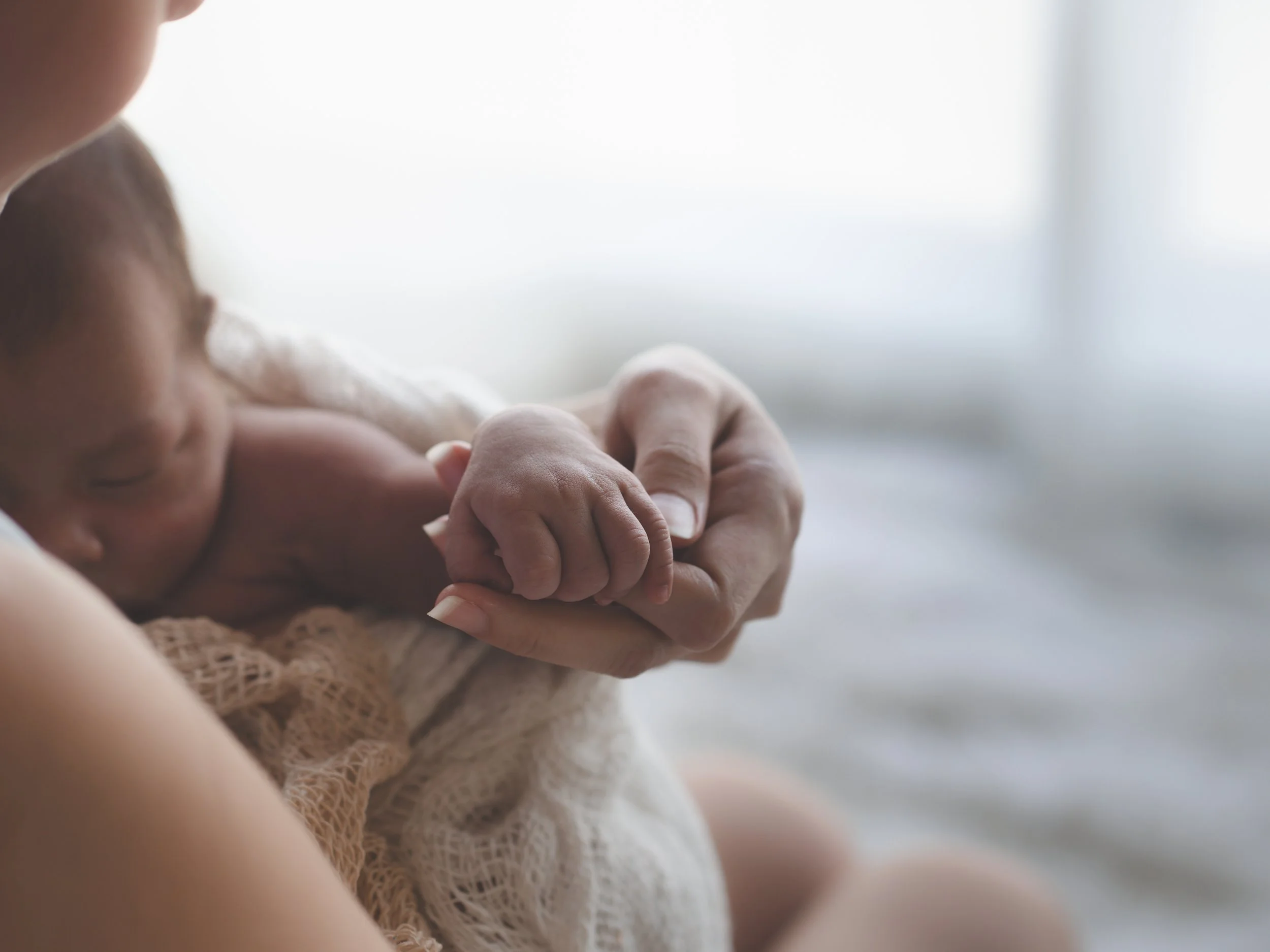 Close-up of a newborn baby holding an adult's finger, with soft lighting and blurred background.