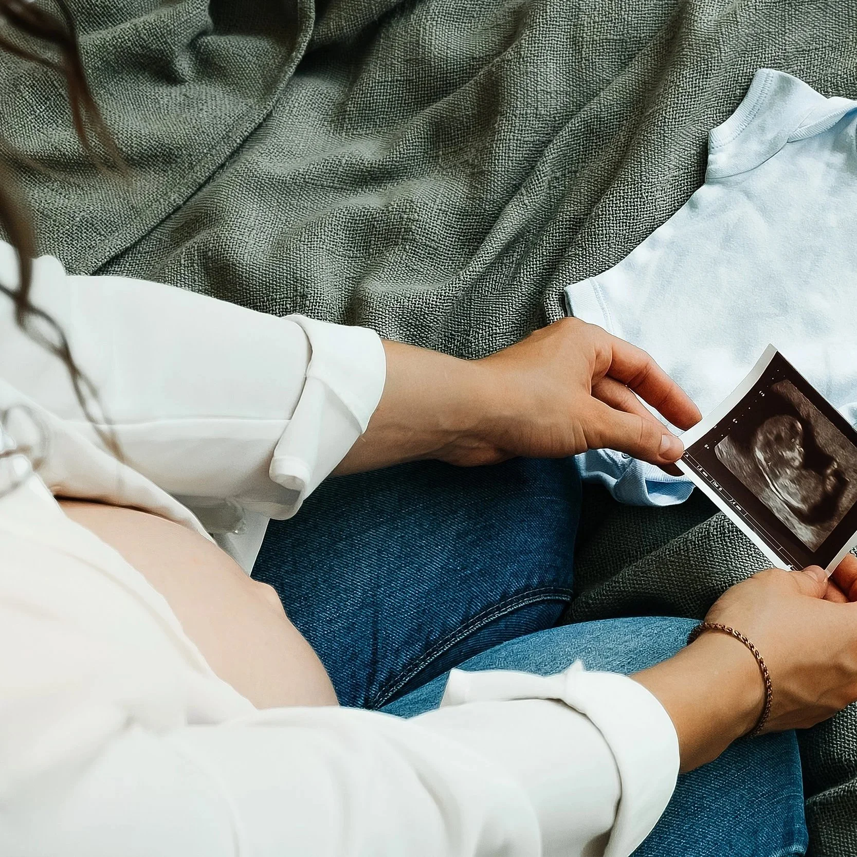 Person holding an ultrasound image, sitting on a couch with a gray blanket, wearing a white shirt and blue jeans.
