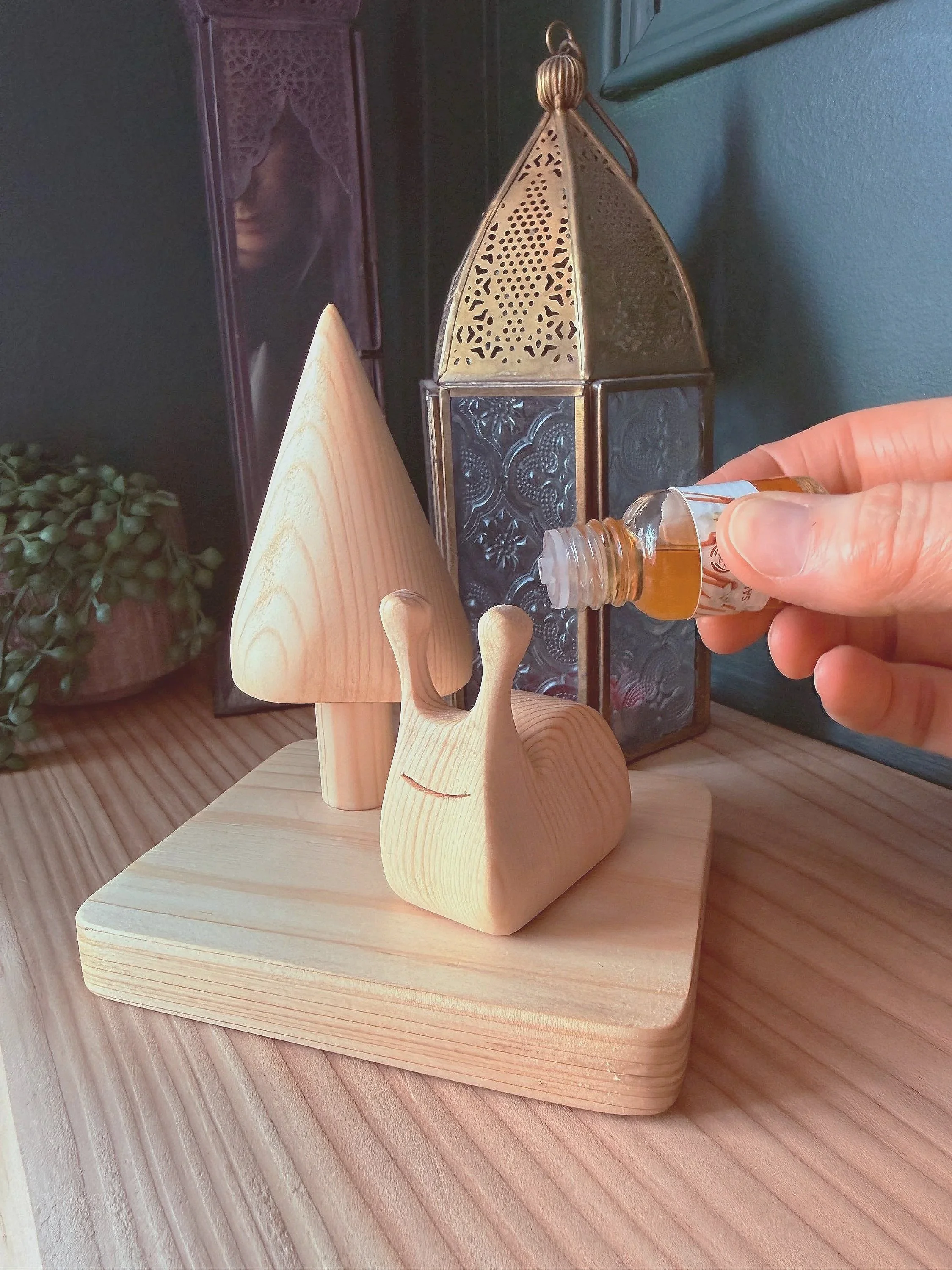 A hand holding a small glass bottle of essential oil over a wooden snail figurine, which is part of a decorative wooden display that includes wooden trees and a brass lantern, on a wooden surface.