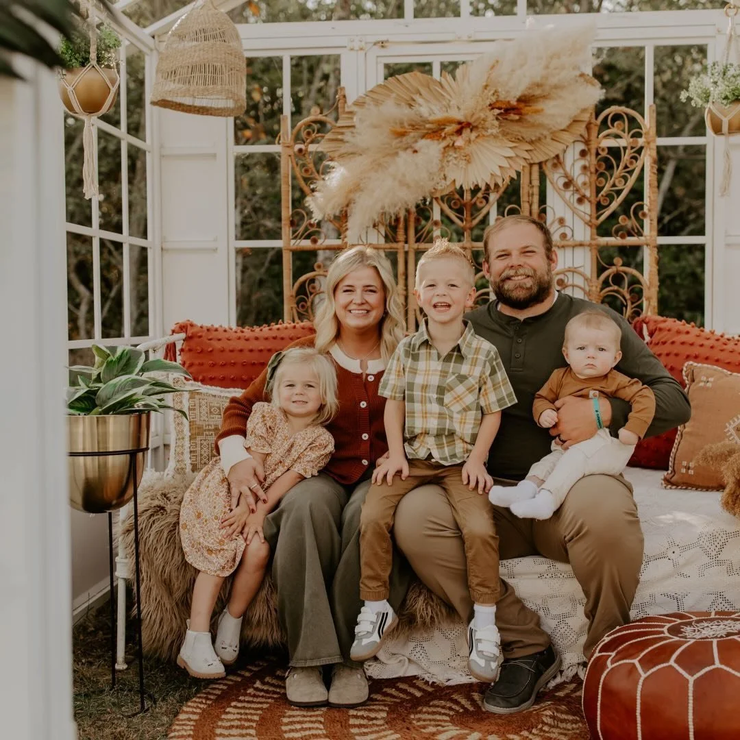 A family of five sitting in a cozy, sunroom with decorative pillows, plants, and window frames, smiling for a portrait.