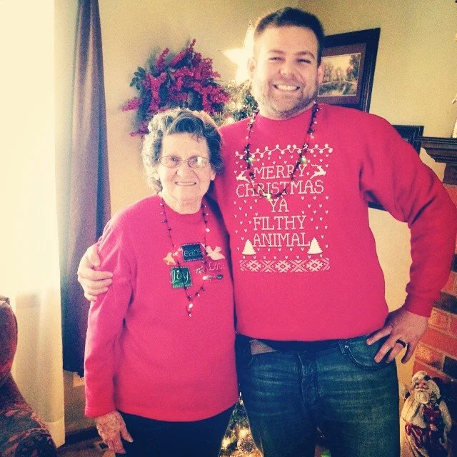 A young man and an elderly woman are standing together indoors, both wearing red Christmas sweaters. The woman’s sweater has holiday decorations and a name tag that says "Joy," while the man's sweater has a Christmas message that reads "Merry Christmas Ya Filthy Animal." They are smiling, and there is a decorated Christmas tree and holiday decorations behind them.