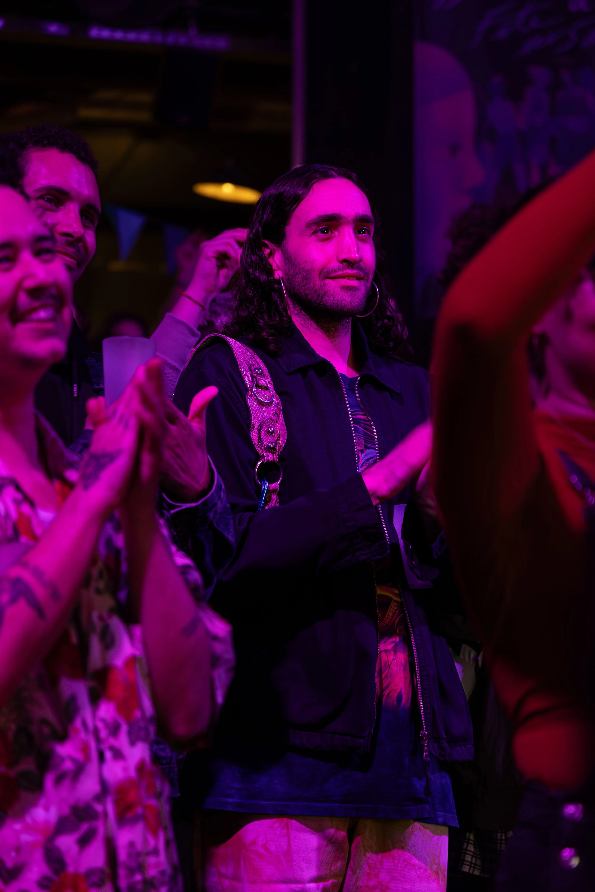 People attending an event, illuminated with purple and pink lighting, clapping and watching something on stage.