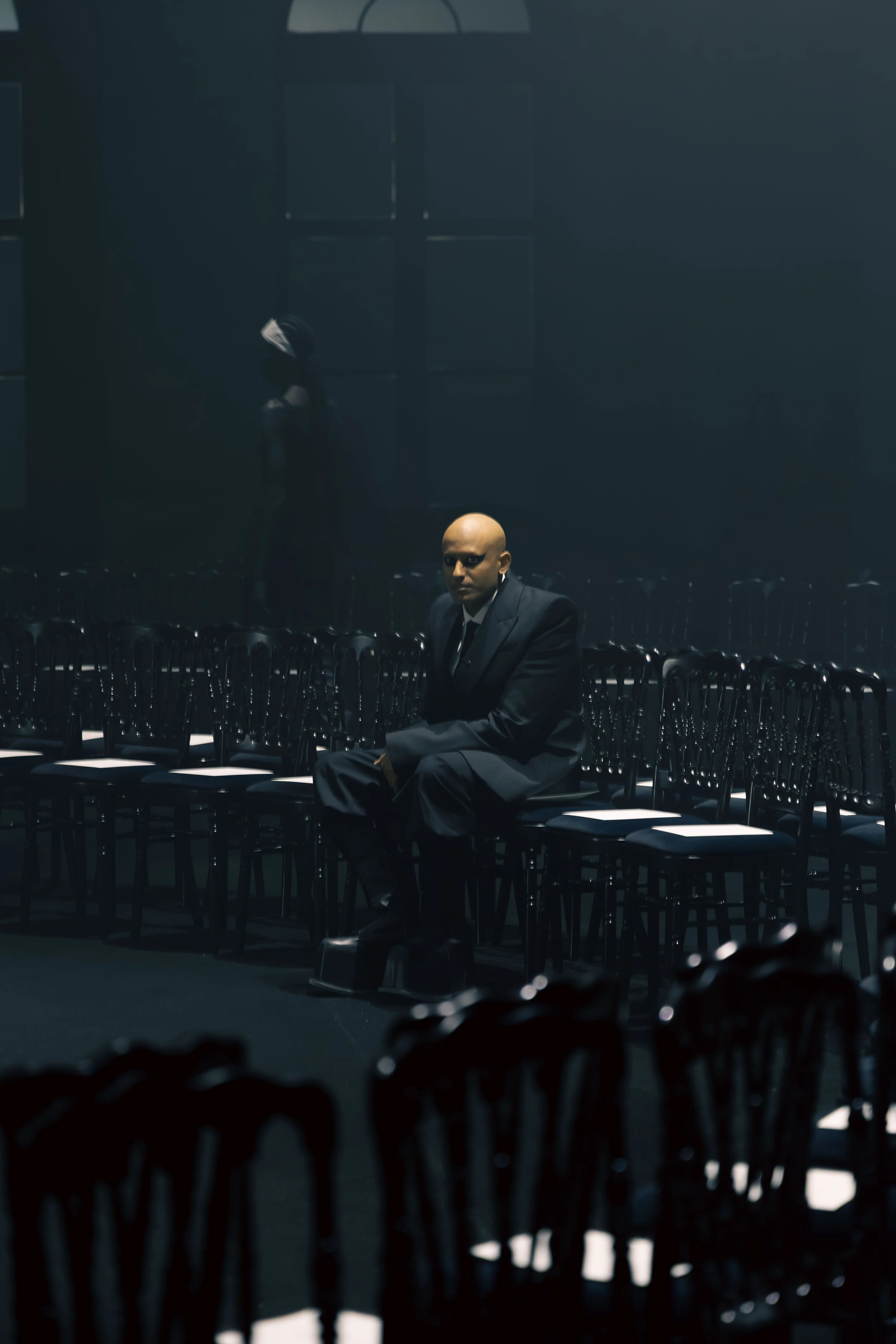 A man in a suit sitting alone in a dark, empty room filled with rows of empty chairs.