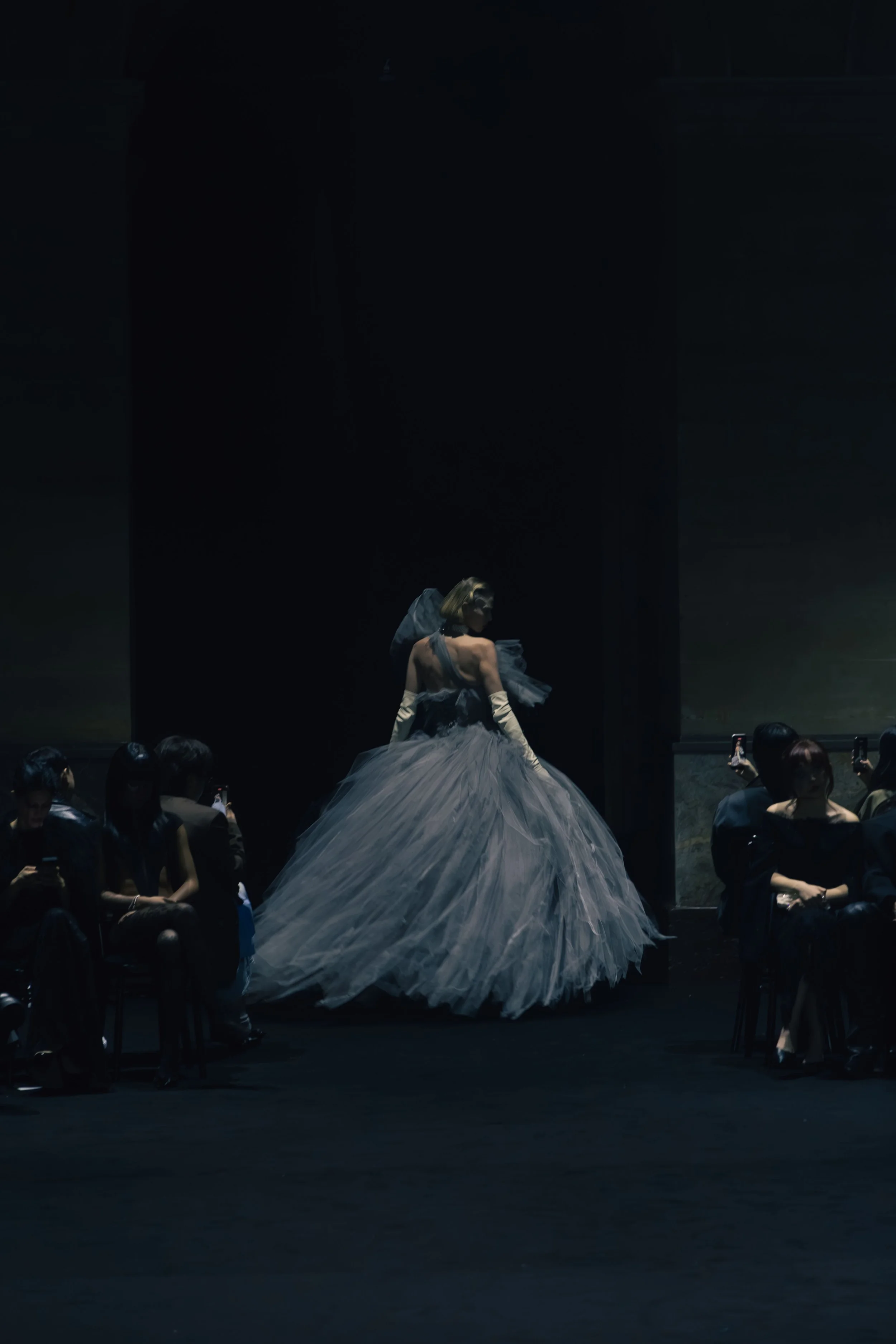 A woman in a dramatic gray gown with a full, voluminous skirt walks on stage during a fashion show, with audience members seated and taking photos.