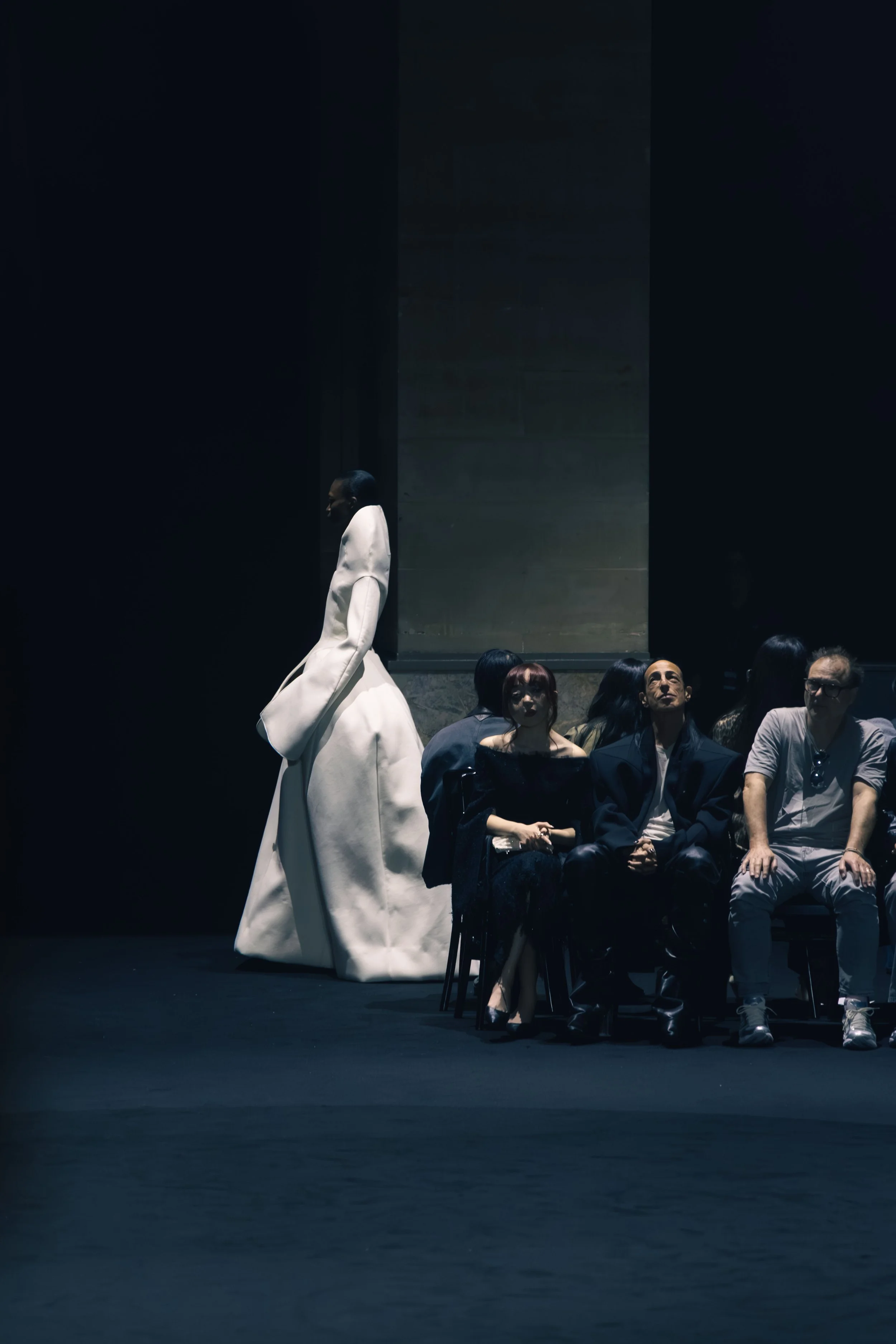 A fashion model in an oversized white coat walking past seated audience members in a dark, minimalist setting.