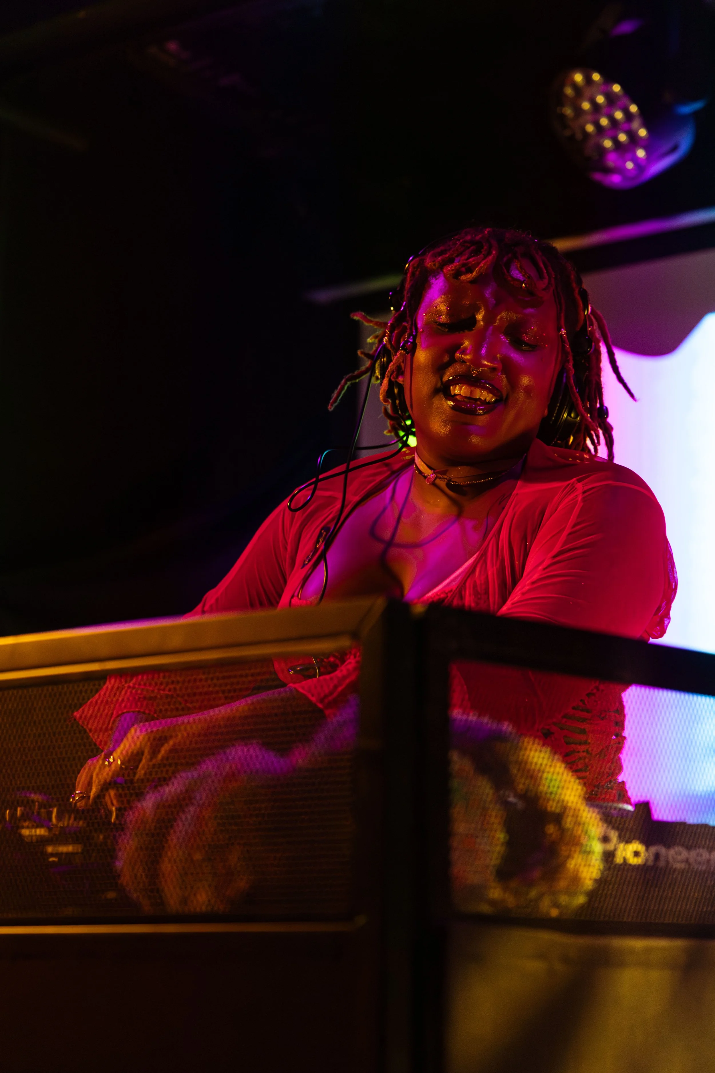 A woman with dreadlocks and a nose piercing, wearing a red top, is djaying in a dimly lit environment with colorful lighting.