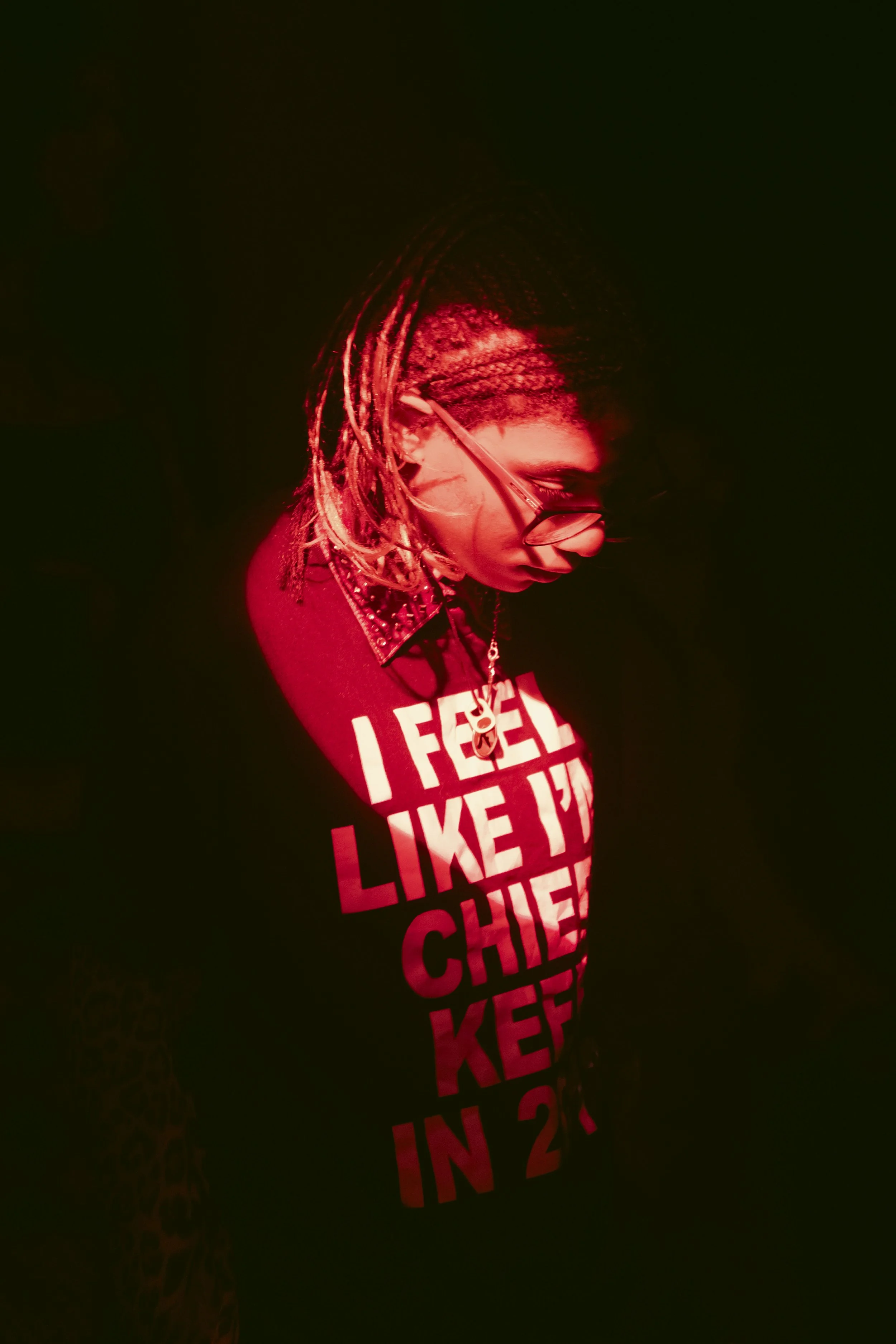 A woman with braided hair and glasses is illuminated by red lighting, wearing a black shirt with bold white and red text, in a dark environment.