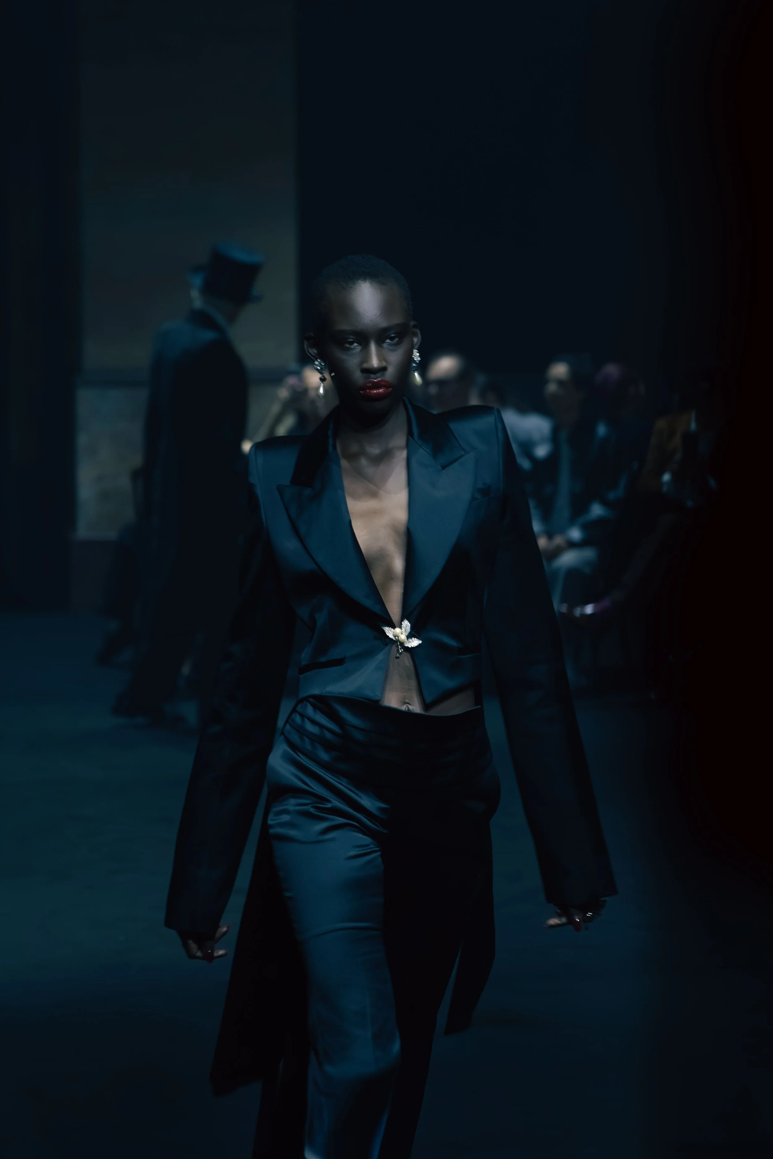 A woman in a black satin blazer and matching pants walking in a dark room with a serious expression, handcuffed, with a man in a black suit and top hat blurred in the background.