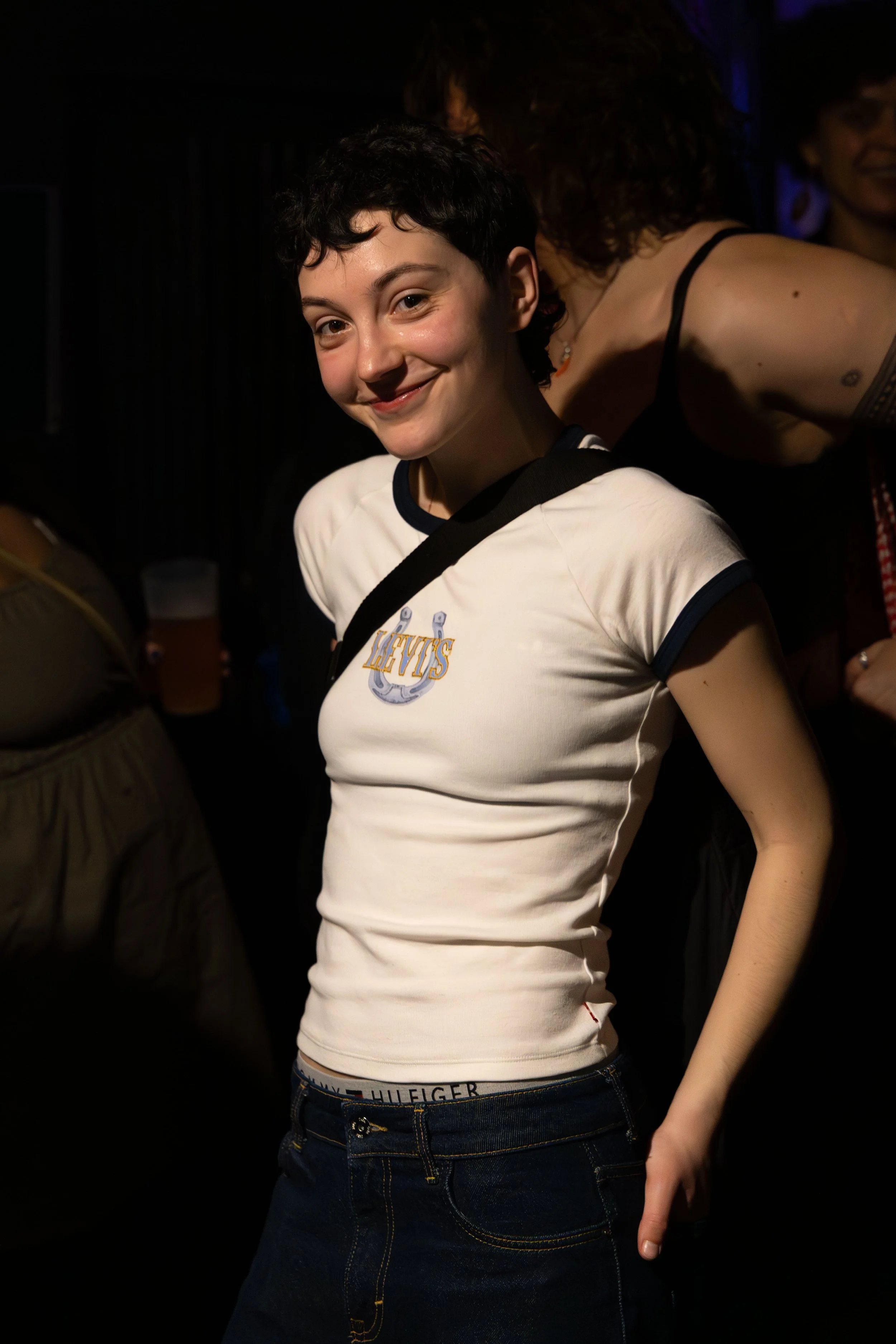 Young woman with short black hair smiling at the camera, wearing a white T-shirt with a nautical anchor logo and dark jeans, with other people in the background in a dimly lit setting.