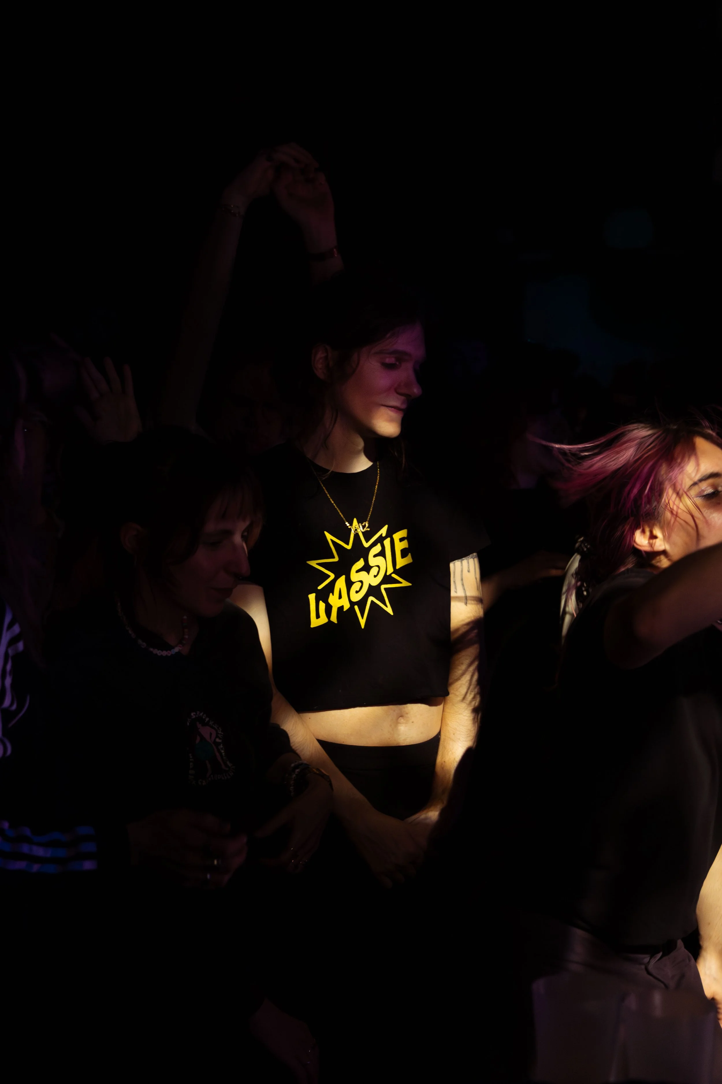 A young woman with dark hair smiling softly, wearing a black T-shirt with the yellow text 'ASSIE' and a star burst graphic, illuminated by colorful stage lighting, in a dark indoor setting with other people.