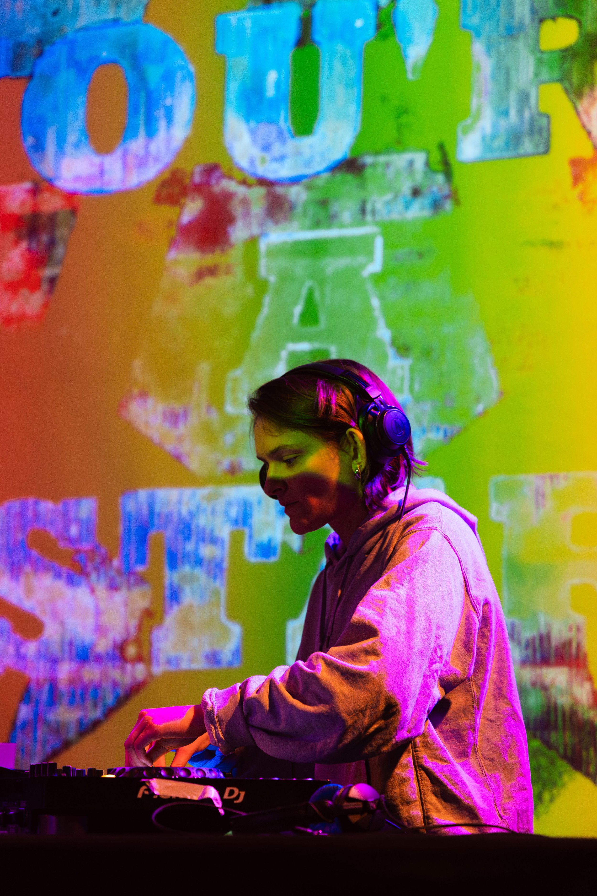 A young person with headphones DJing at a colorful, illuminated event or club, with vibrant abstract background artwork.