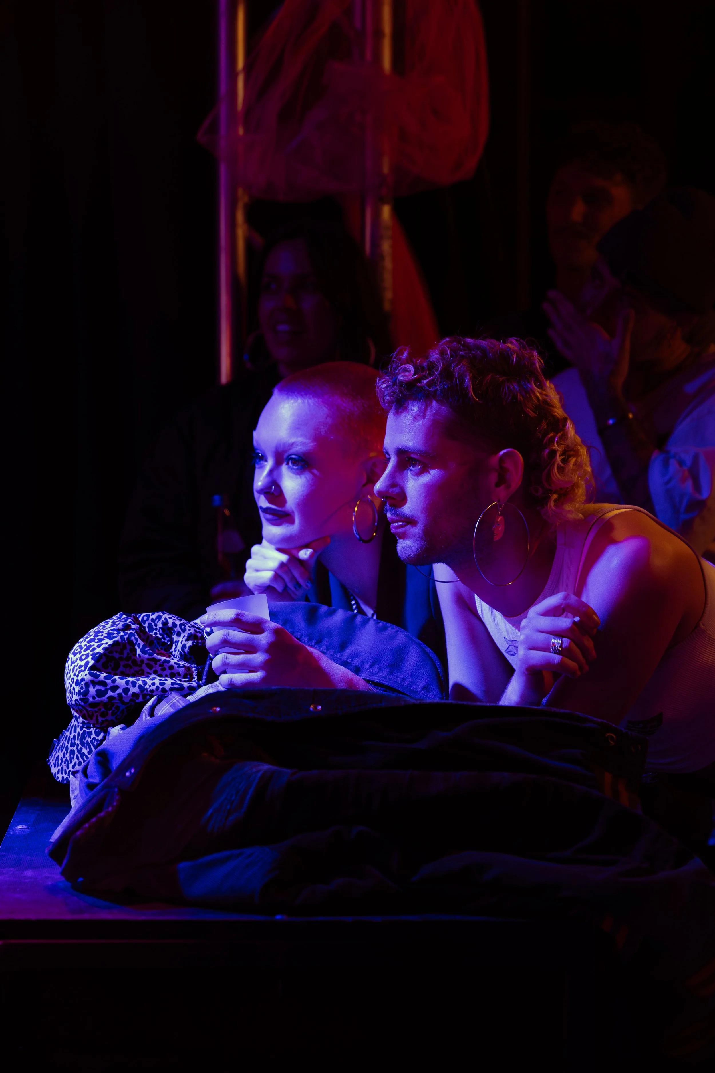 Two individuals sitting at a table in a dark, colorful environment, engaged in conversation or focused on something in front of them, illuminated by vibrant neon lighting.