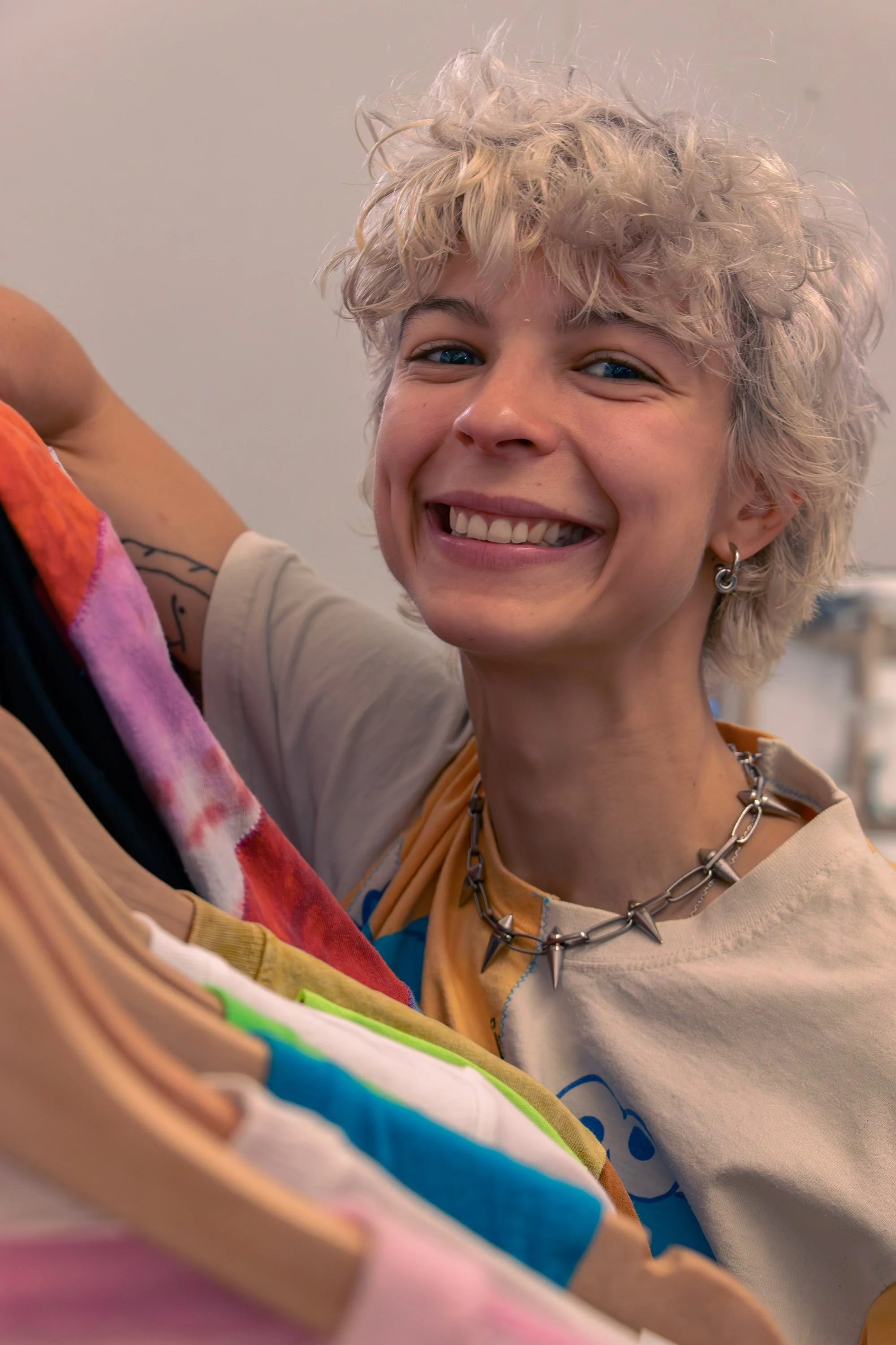 A person with curly, platinum blonde hair smiling at the camera with her colorful clothes on a rack.