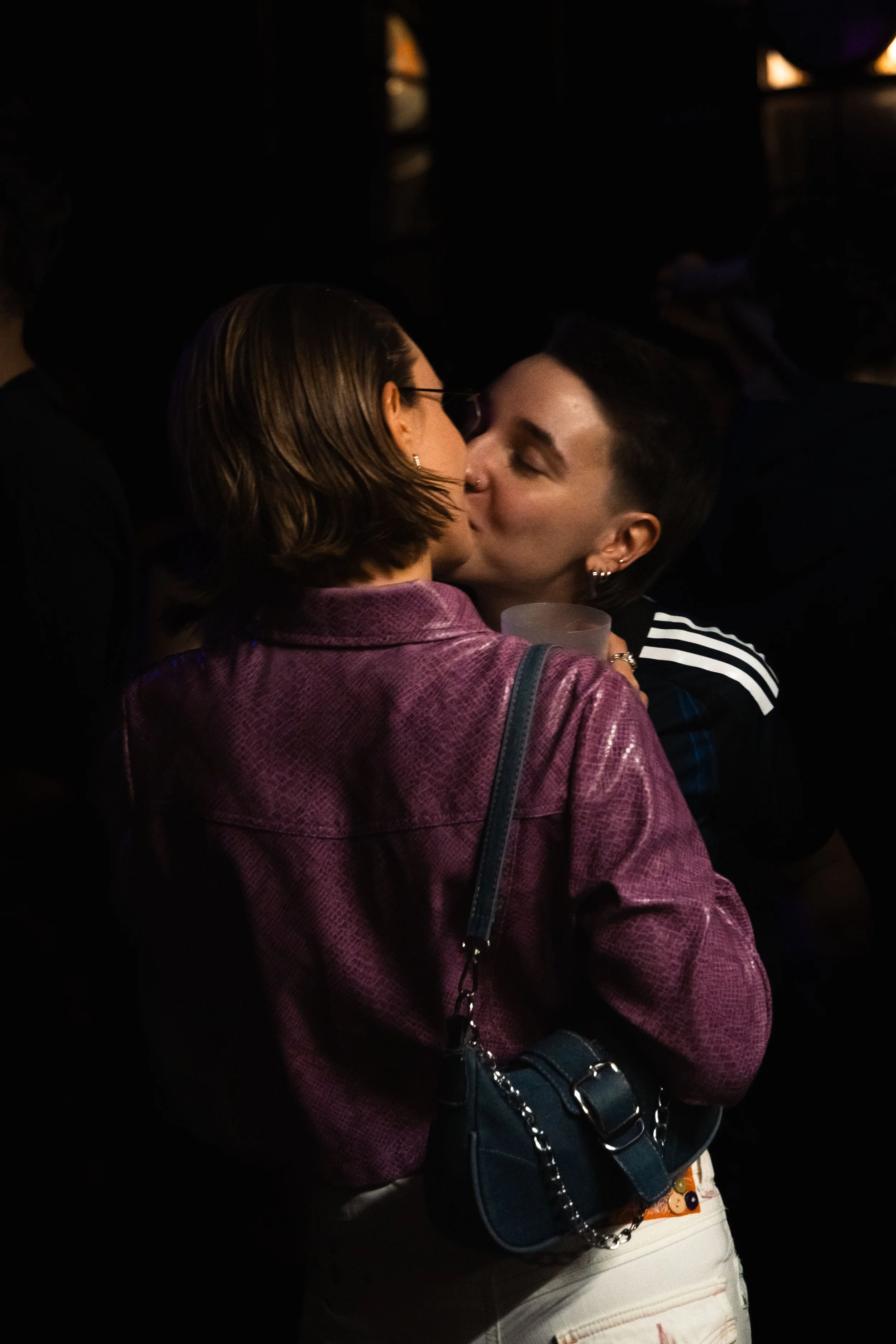 Two women sharing a kiss in a dark environment, one wearing a purple jacket and the other wearing a black shirt with white stripes, carrying a black purse with a chain strap.