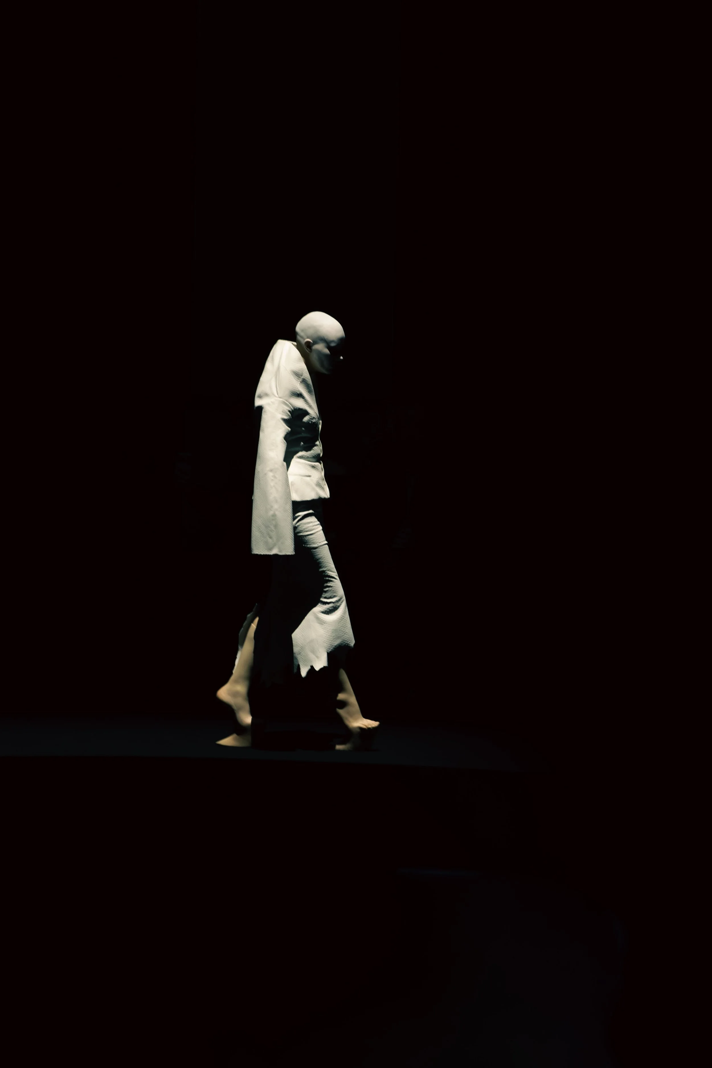 A person wearing a white suit and mask walks in darkness with focused lighting.