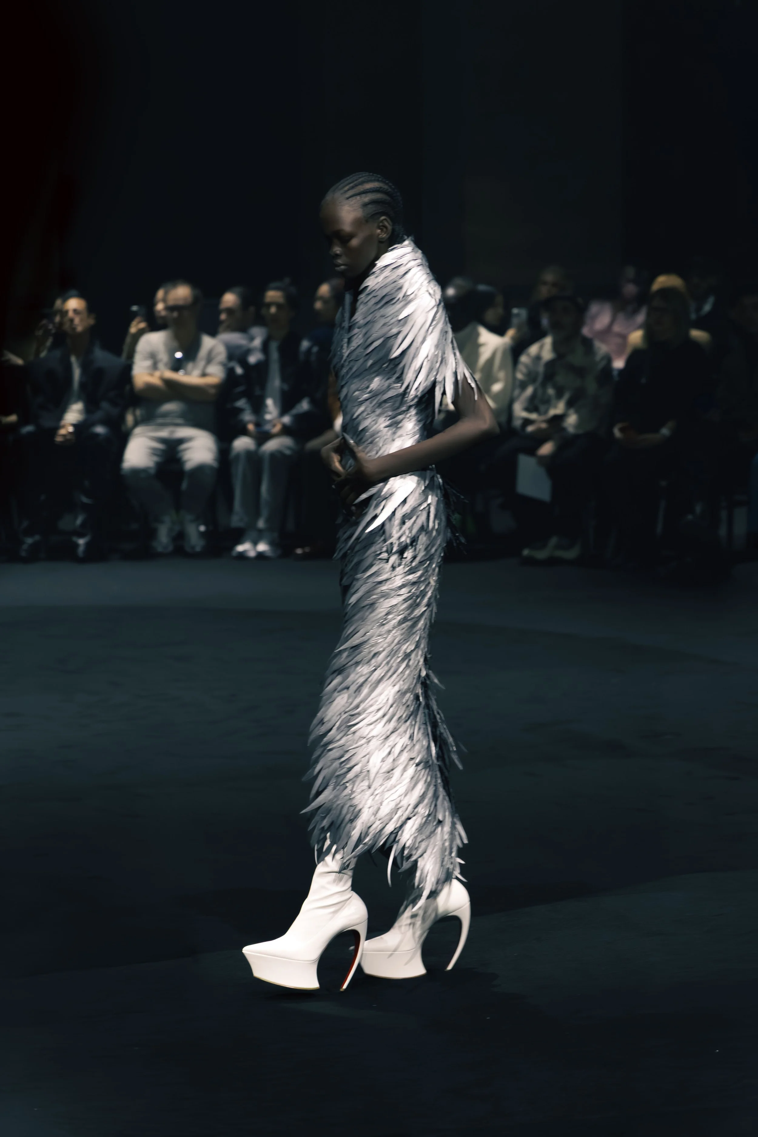 Fashion model walking on runway wearing high white platform shoes and a feathered dress, audience observing in the background.