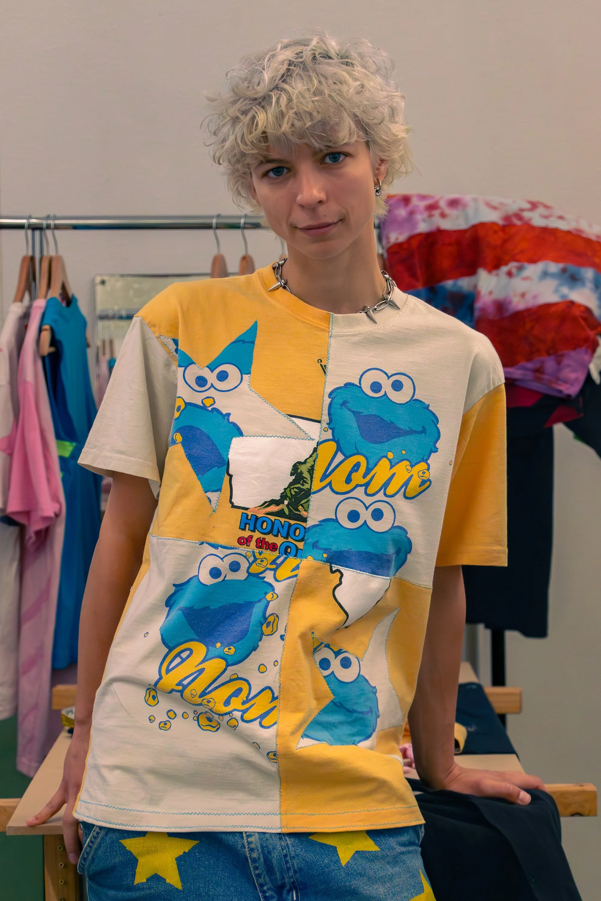 Person with curly blonde hair wearing a colorful T-shirt featuring Sesame Street characters and blue jeans with yellow stars, standing indoors in front of a clothing rack and colorful blanket.