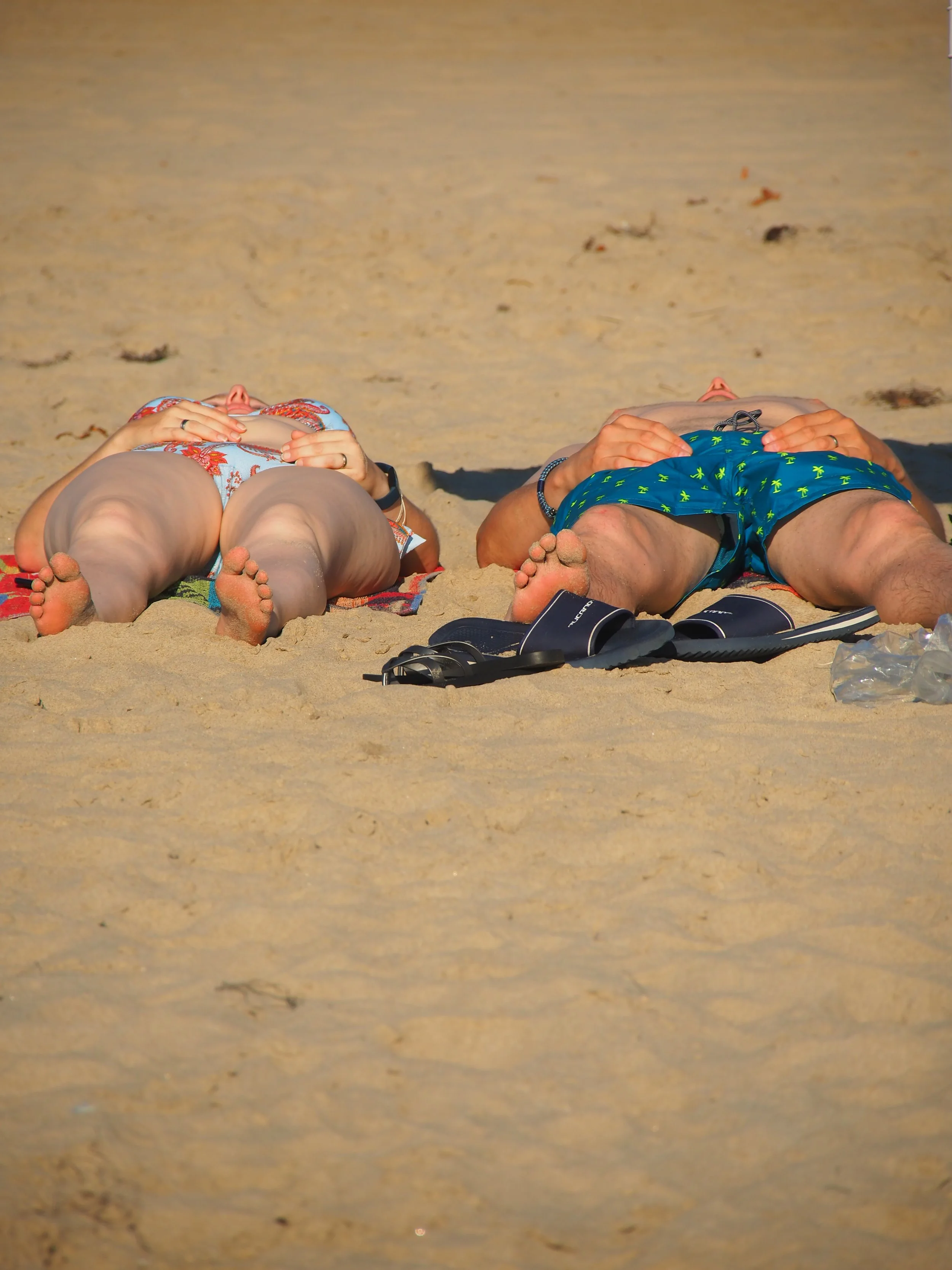 Two people lying on their backs on the sandy beach, facing upwards with their hands on their tummies, wearing swimsuits, with a sandy shoreline in the background.
