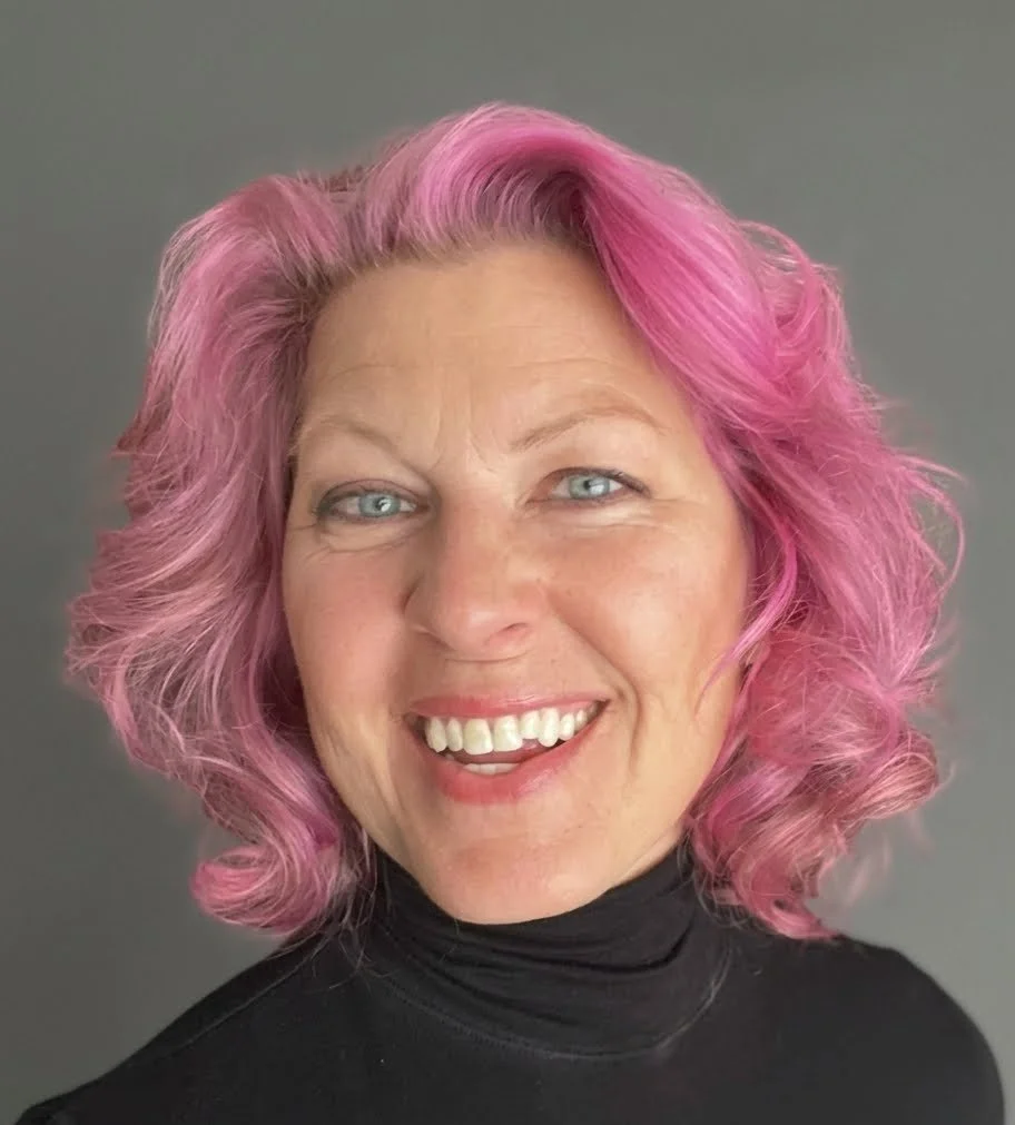 A woman with short, wavy pink hair, blue eyes, and a broad smile, wearing a black turtleneck against a gray background.